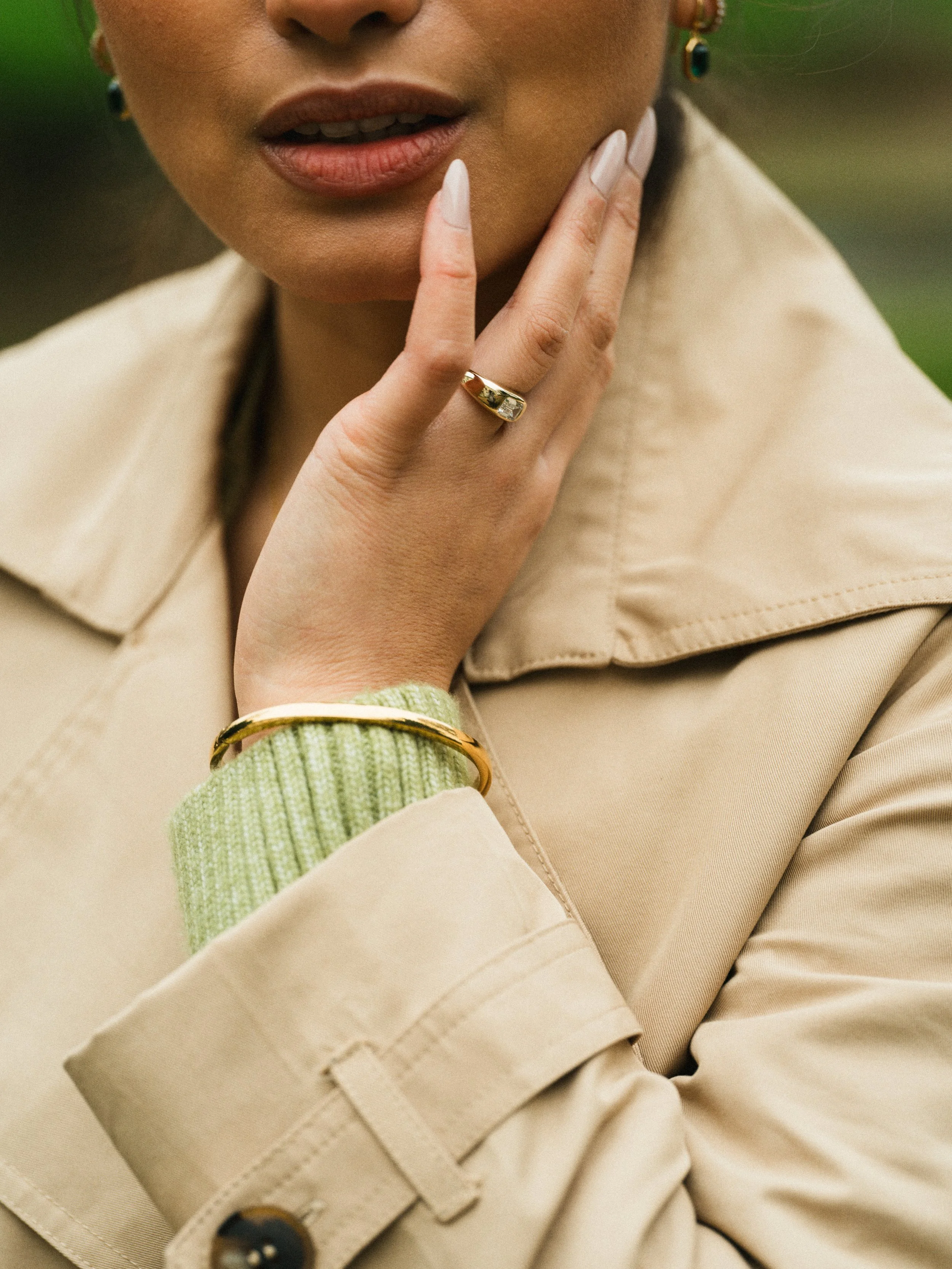 Close-up of woman wearing jewelry, including a ring, bracelet, and earrings, with her hand touching her chin and lips, dressed in a beige trench coat and light green knit sweater.