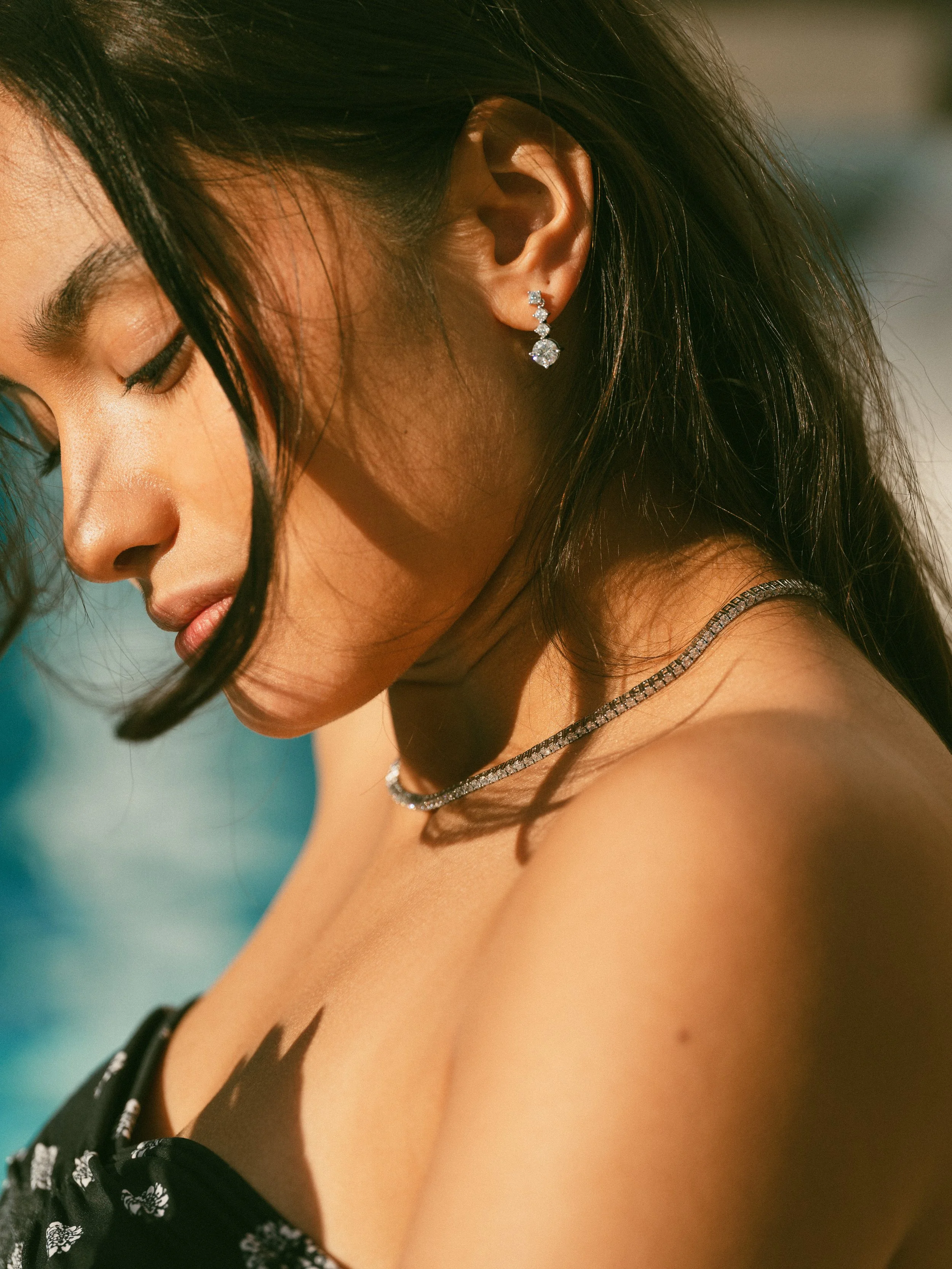 A woman with dark hair and earrings, wearing a necklace and a black top with white floral patterns, looking downward with closed eyes, on a sunny day.