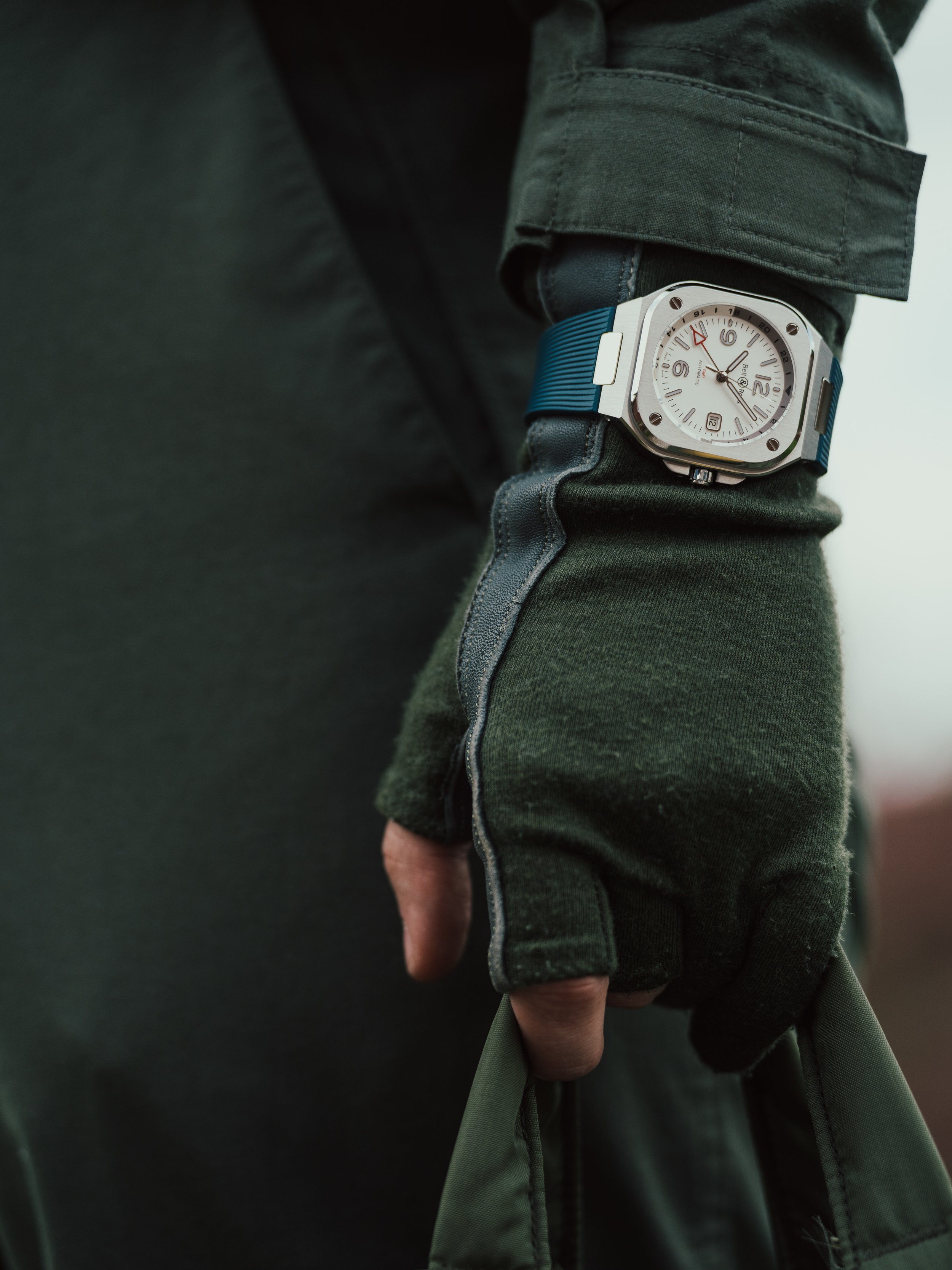 Close-up of a person's wrist wearing a silver watch with a white face, black numerals, and a blue band, partially covered by a dark green jacket sleeve. The person is also wearing a dark green glove under the jacket, with their hand gripping a green 