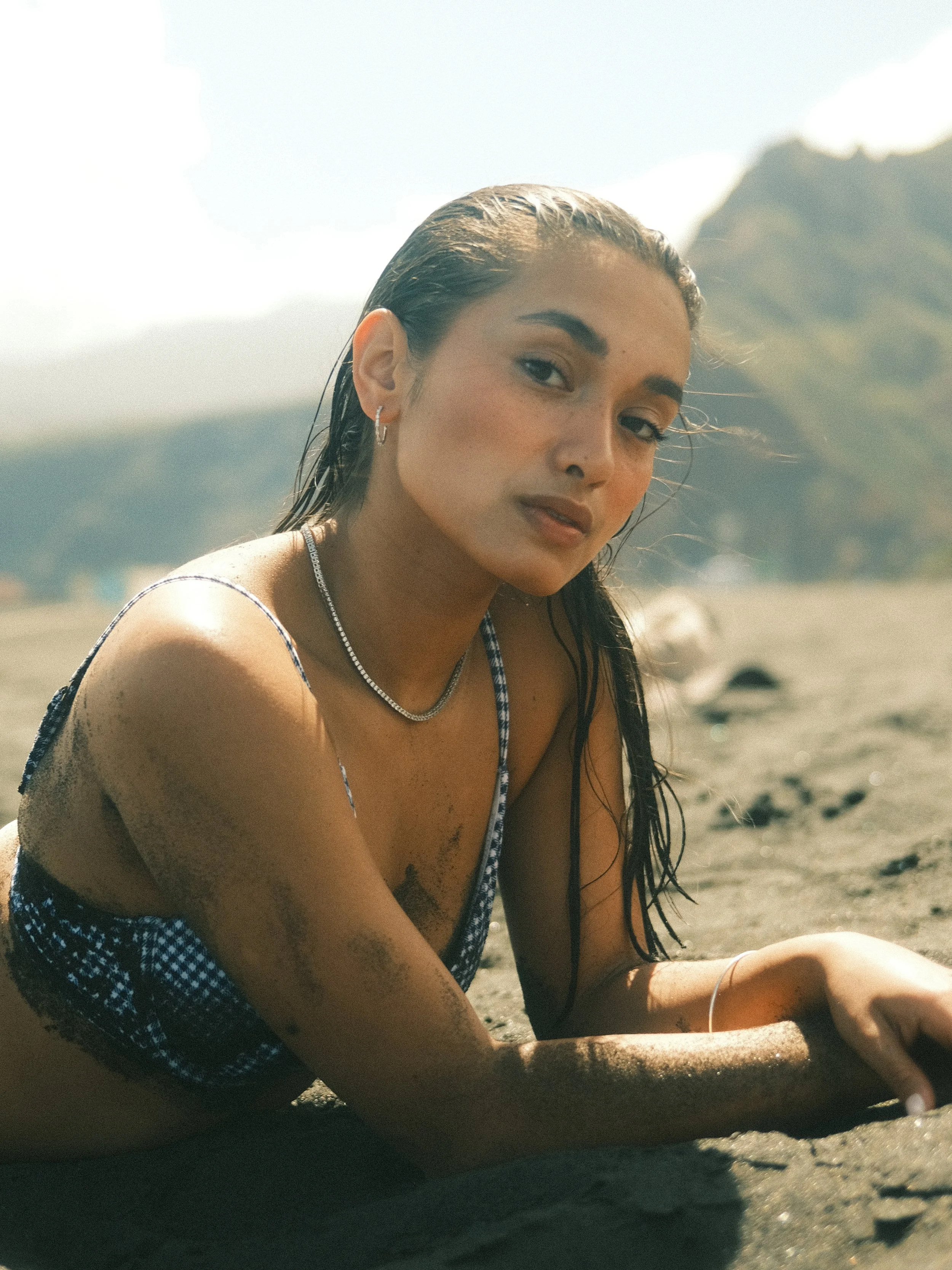 A young woman lying on a sandy beach, wearing a blue checkered bikini and jewelry, with sandy hair and a mountain in the background.