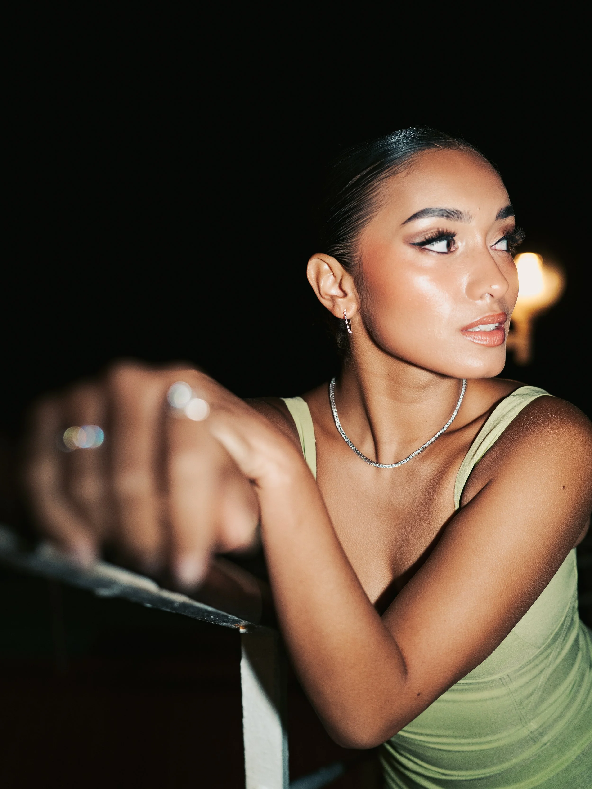 A young woman with dark hair slicked back, wearing a lime green dress, jewelry, and makeup, is gazing to the side with a serious expression, leaning on a railing at night.