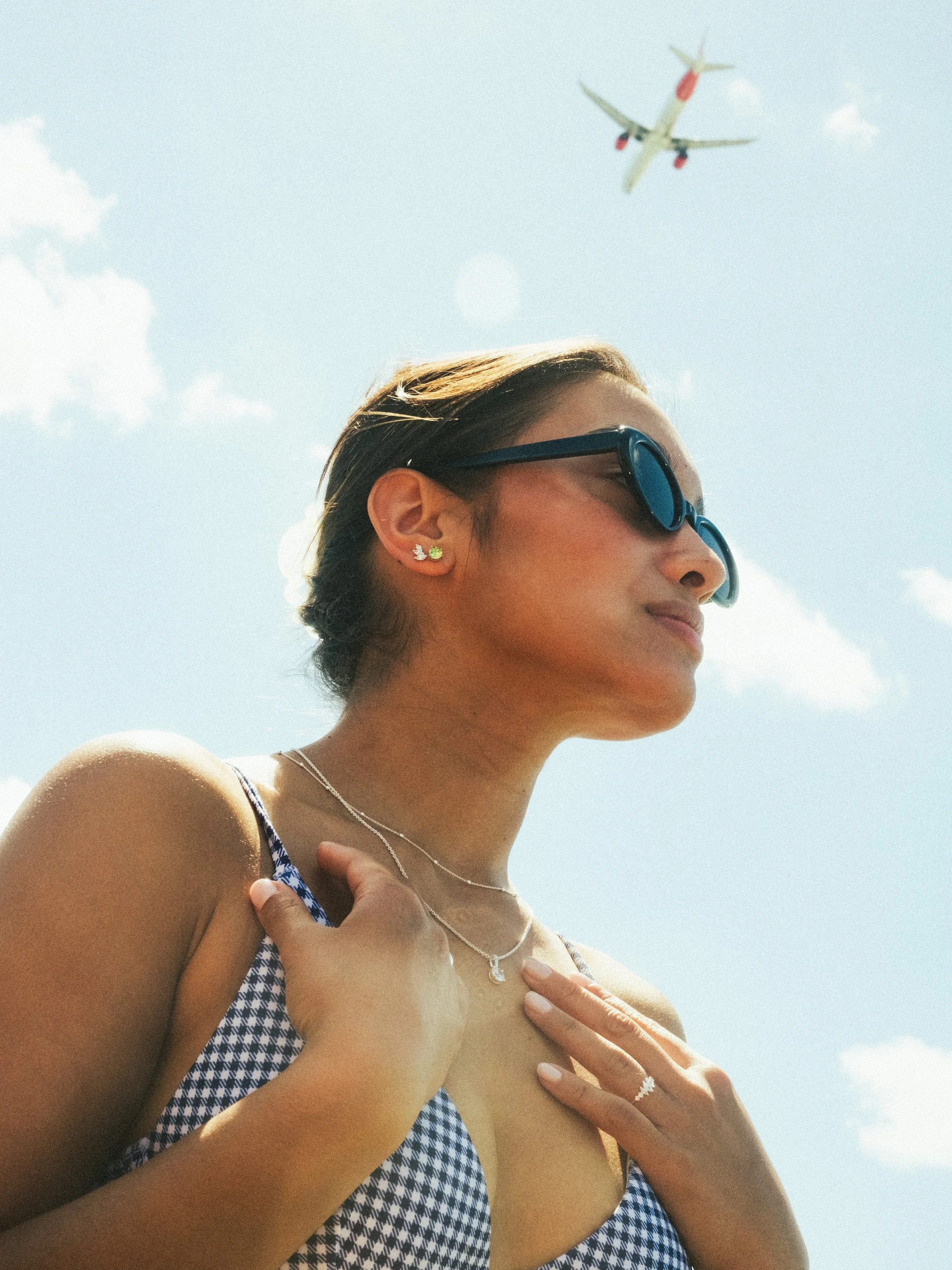 A woman wearing sunglasses and jewelry standing outdoors, with an airplane flying overhead in a clear, partly cloudy sky.
