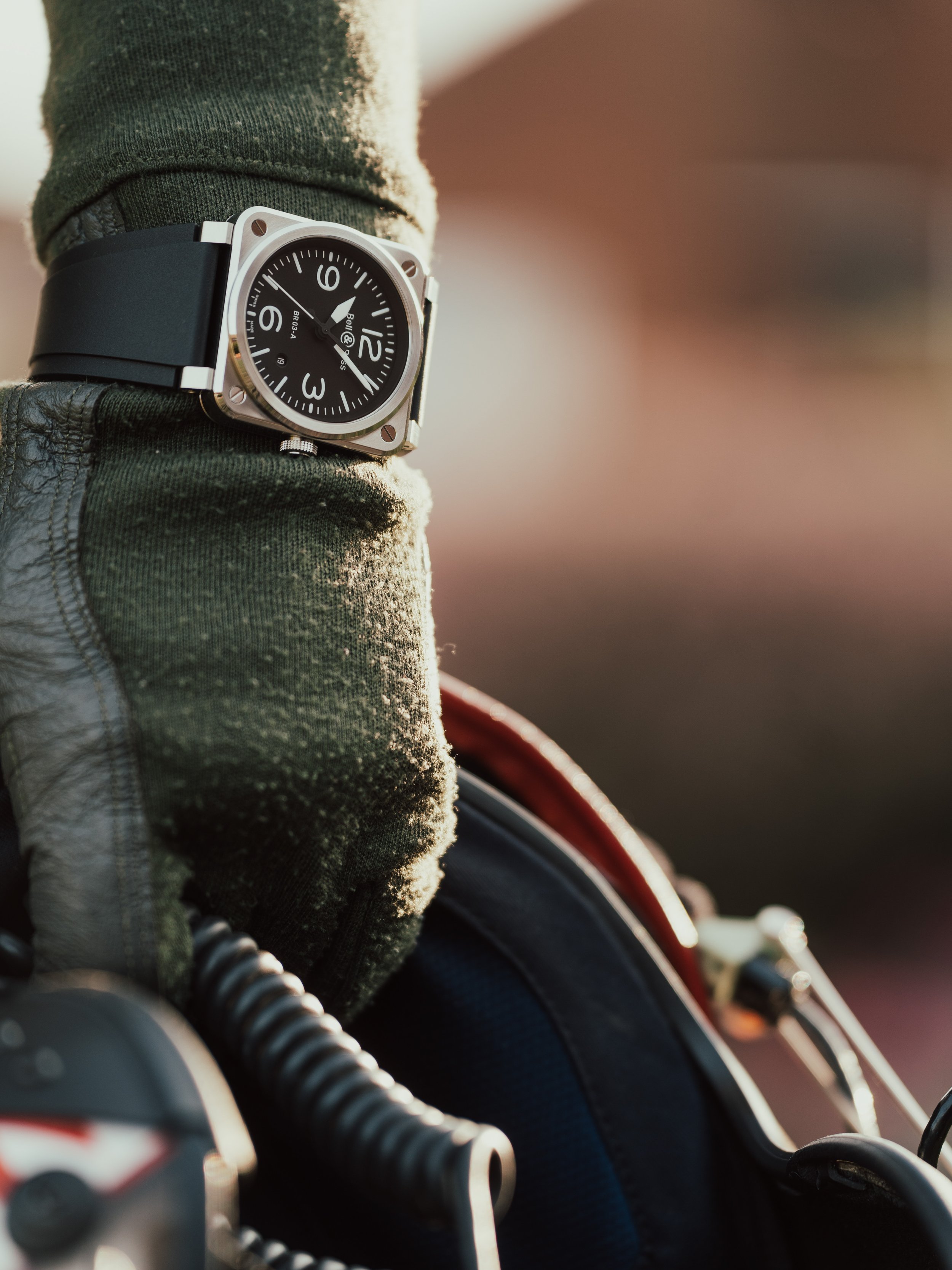 Close-up of a person's wrist wearing a square watch with a black dial, white numbers, and a black strap, as the person grips a motorcycle handlebar wearing a textured glove, with a blurred background.