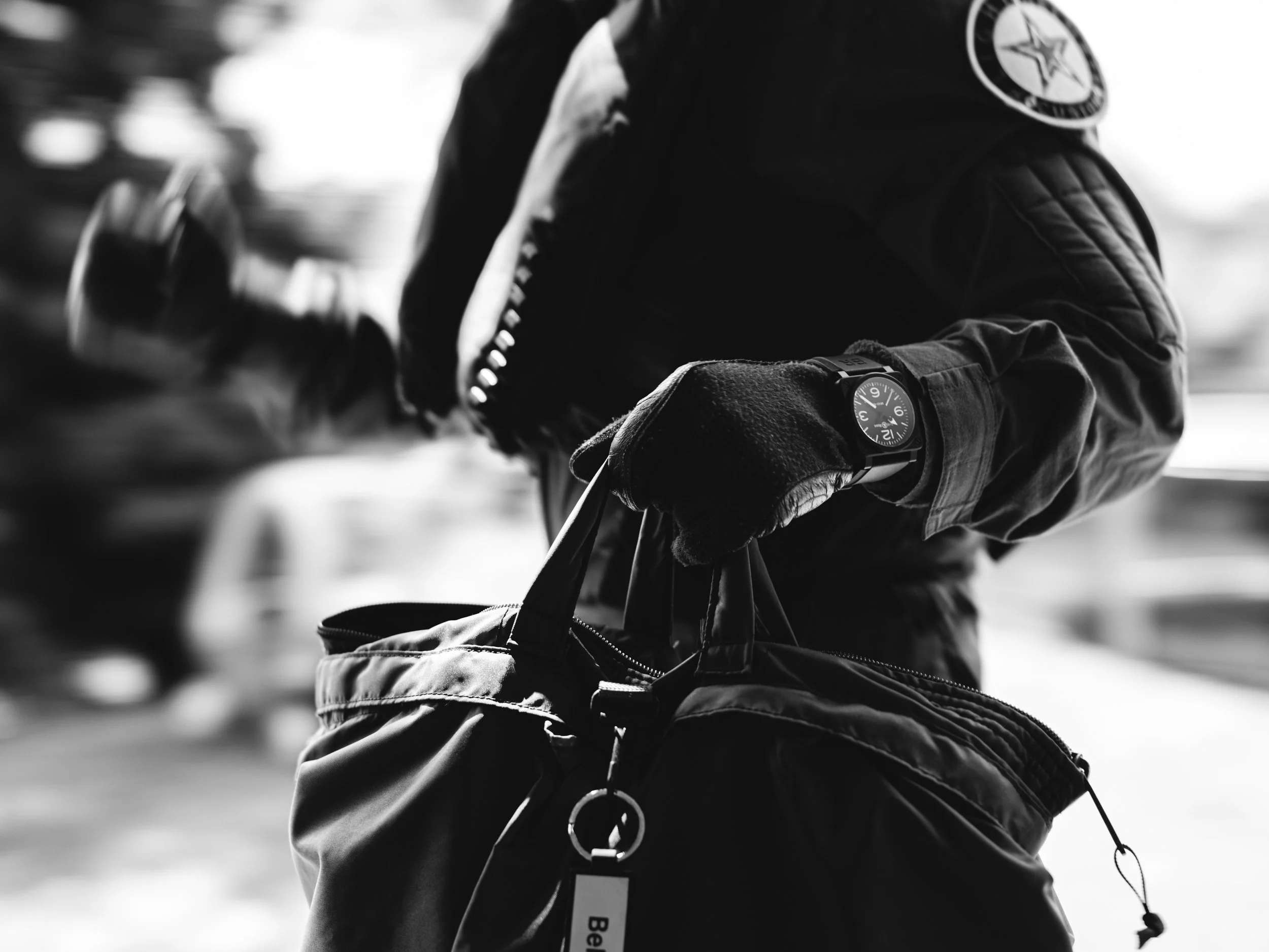 Close-up of a person wearing a jacket, gloves, and a watch, holding a bag, with a blurred background.