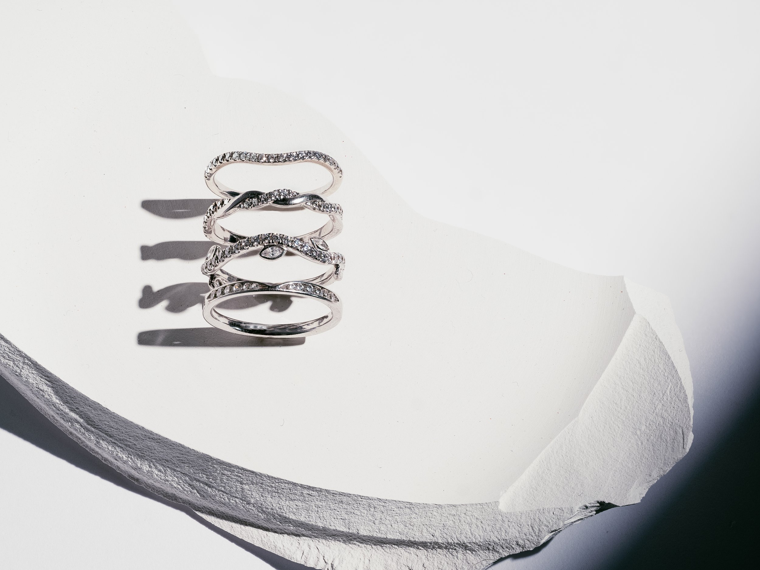 Four silver rings with diamonds stacked on white textured surface with torn paper edge casting shadows.