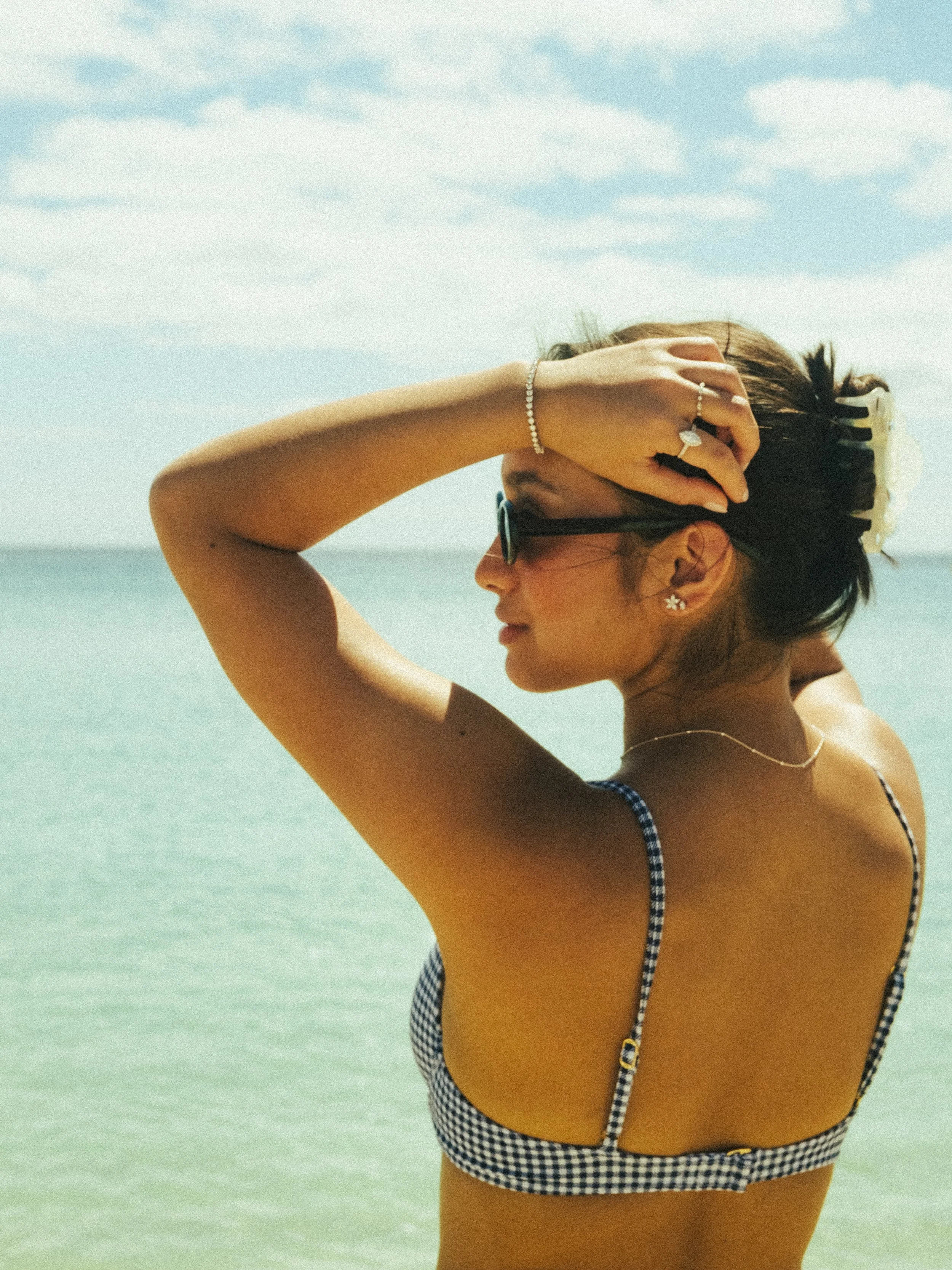 Woman at the beach wearing sunglasses and a checkered bikini, with her hand on her head, holding a hair clip, and looking to the side.