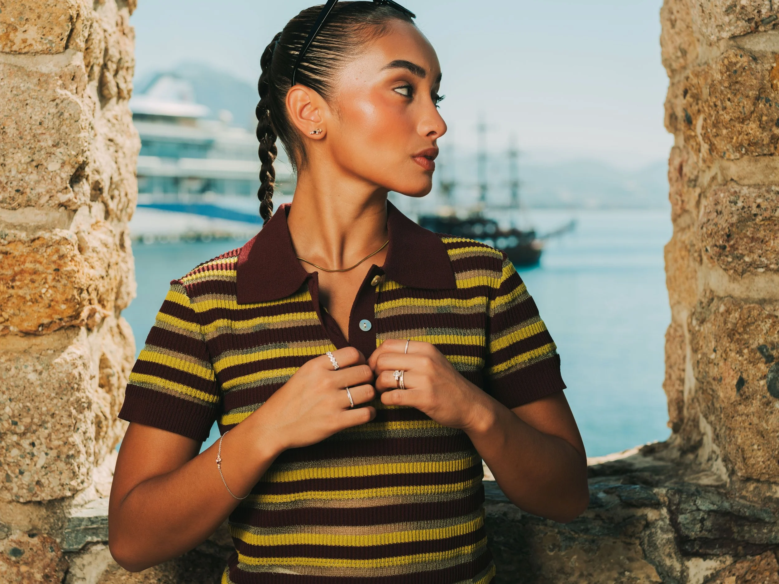 A woman with slicked-back hair in a braid, wearing a striped polo shirt and jewelry, stands between stone walls near water with boats and a ship in the background.