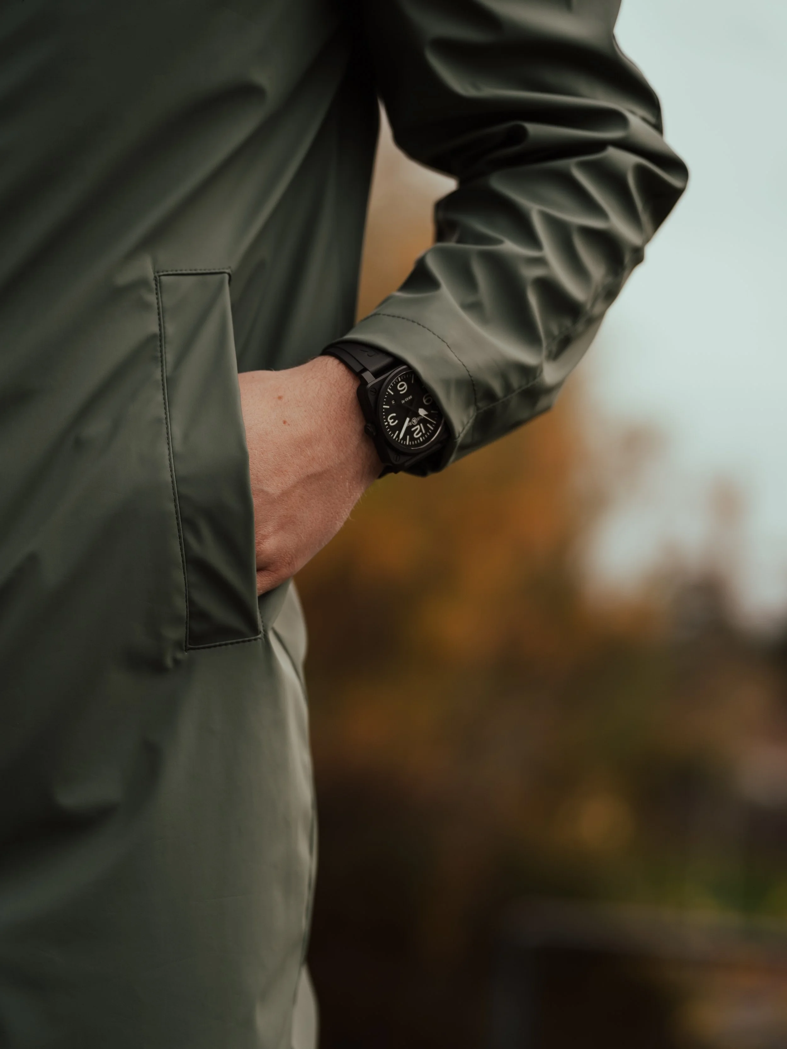 Close-up of a person's wrist wearing a black watch with a large round dial, in a green jacket with their hand in the jacket pocket, outdoors with blurred autumn foliage in the background.
