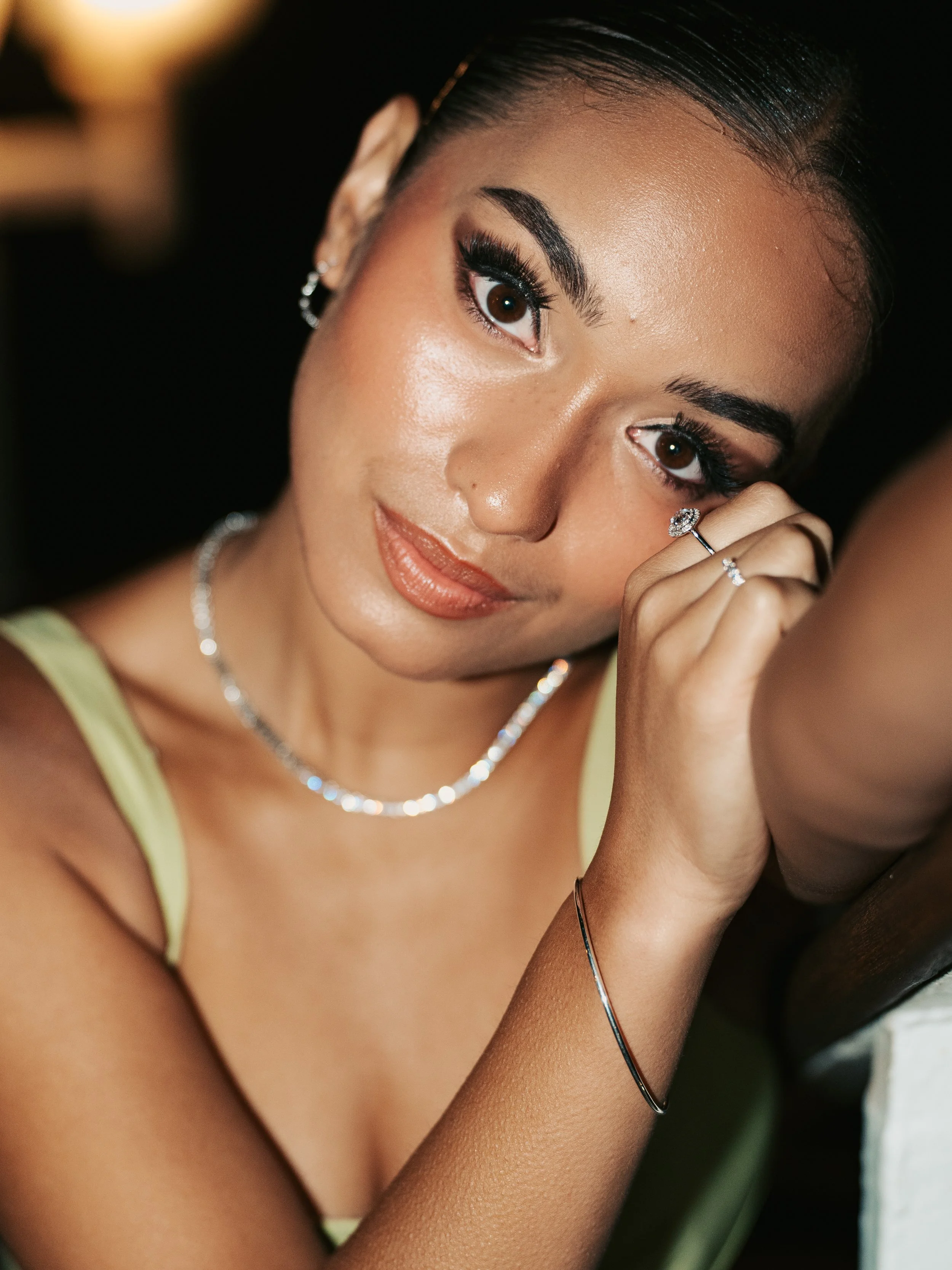 A woman with dark hair and makeup, wearing jewelry, and touching her face with one hand while looking at the camera.