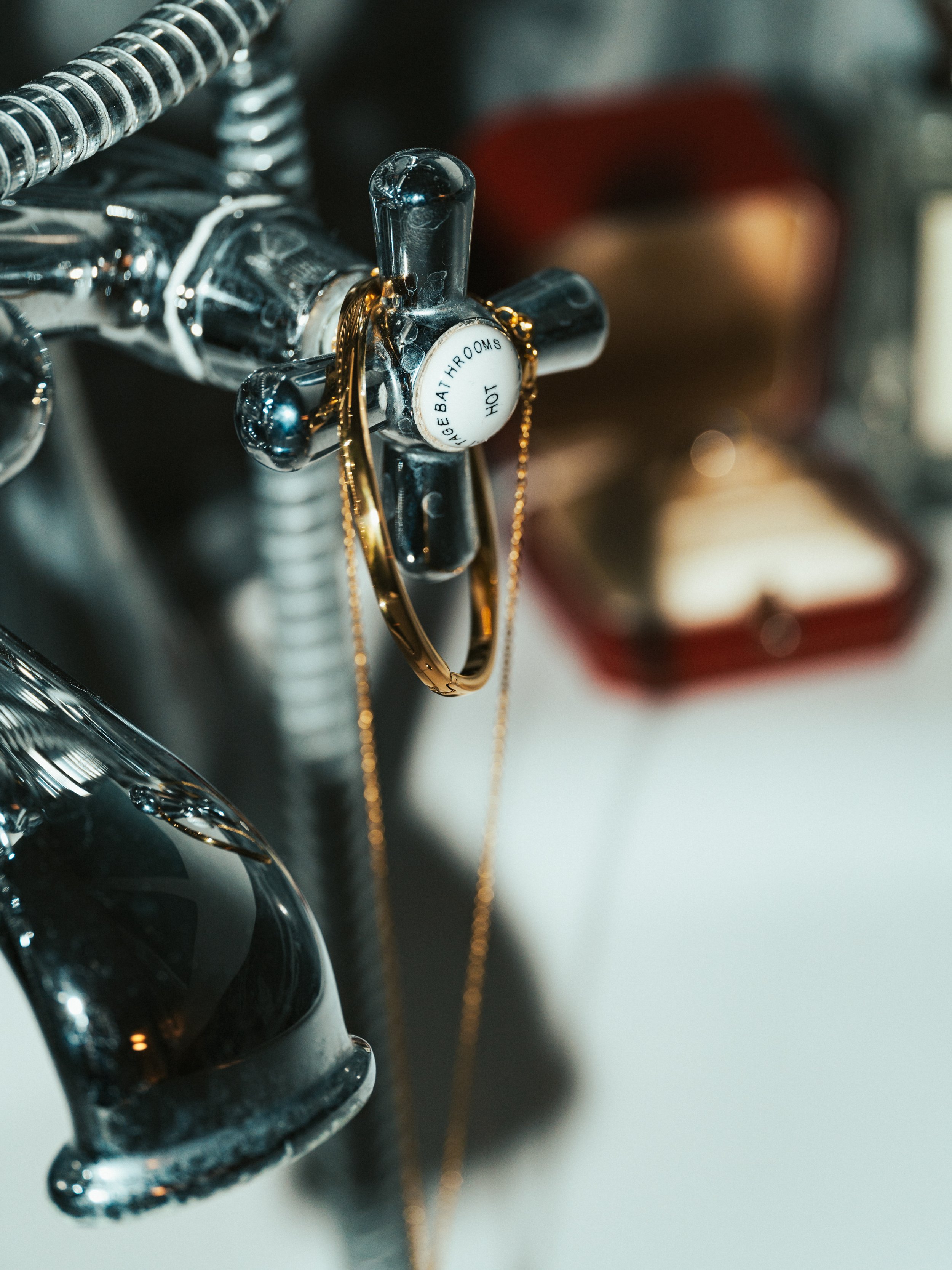 Close-up of a faucet with a white knob labeled 'HOT, BEAT BOYS, BATHROOMS'. Gold jewelry is hanging on it, with a blurry red box in the background.