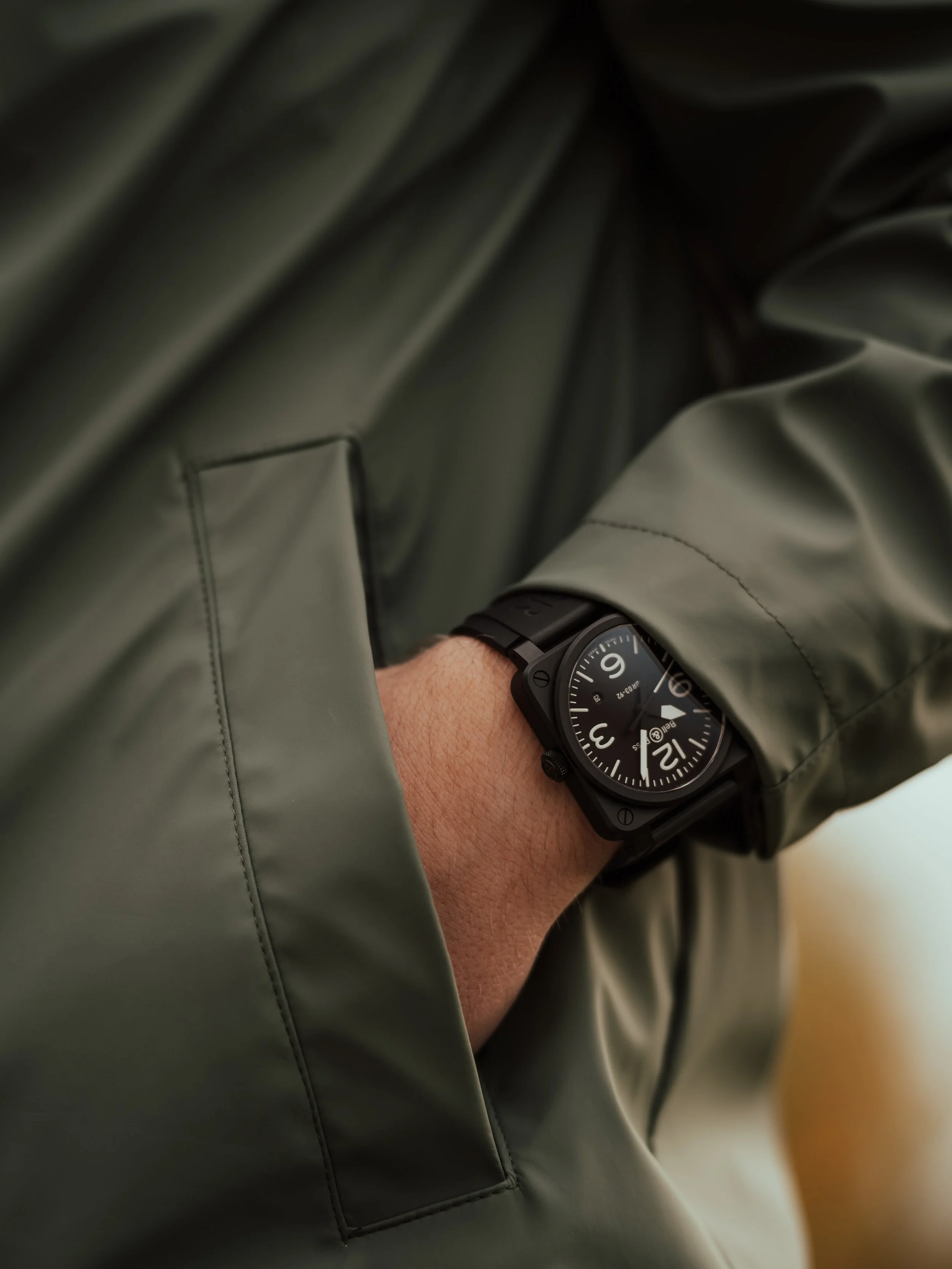 A person's arm resting in the pocket of a dark green jacket, wearing a black wristwatch with a round face and large white numbers.
