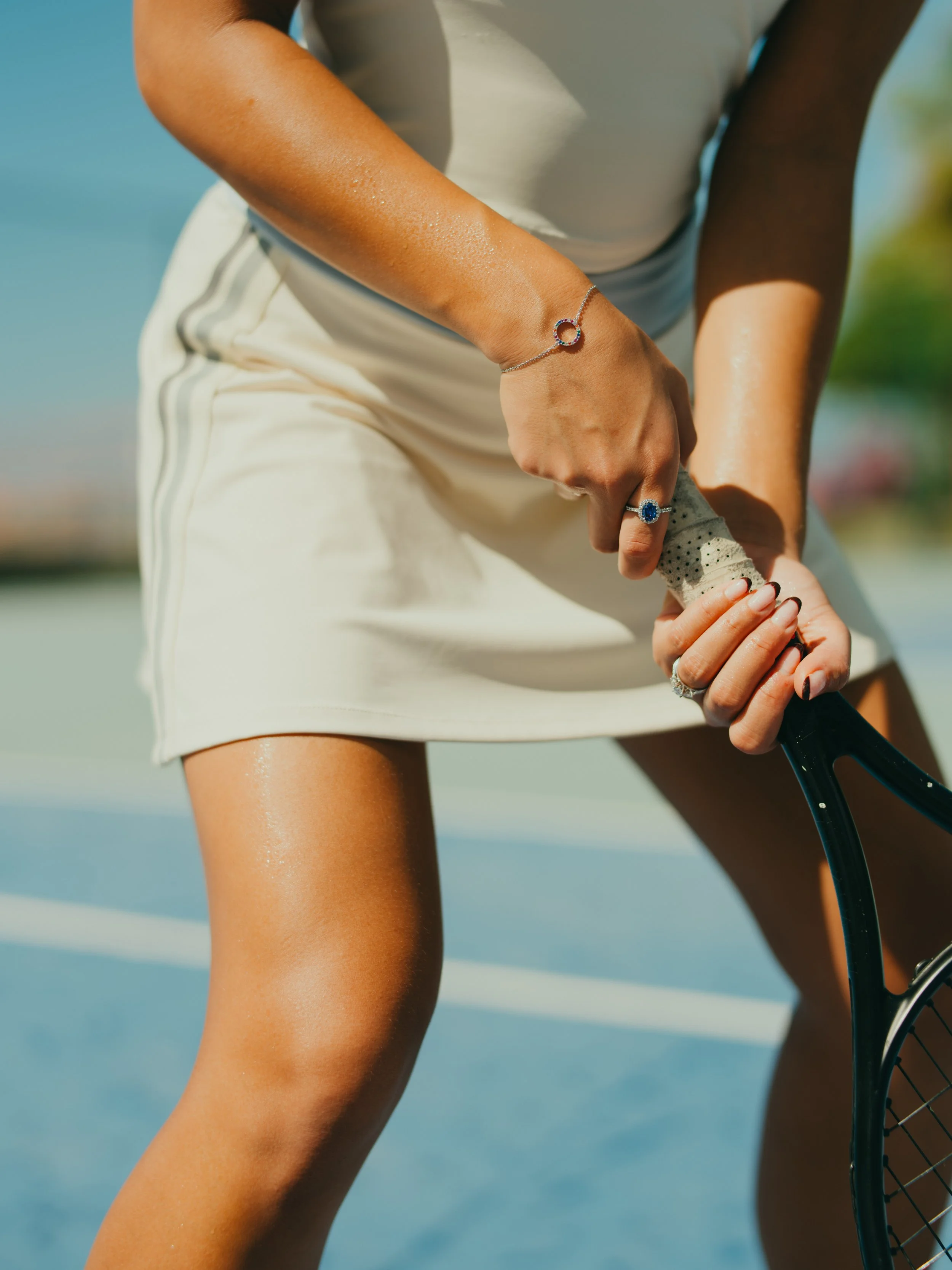 Close-up of a woman wearing a white sports dress and jewelry, gripping a tennis racket on a tennis court during sunny weather.