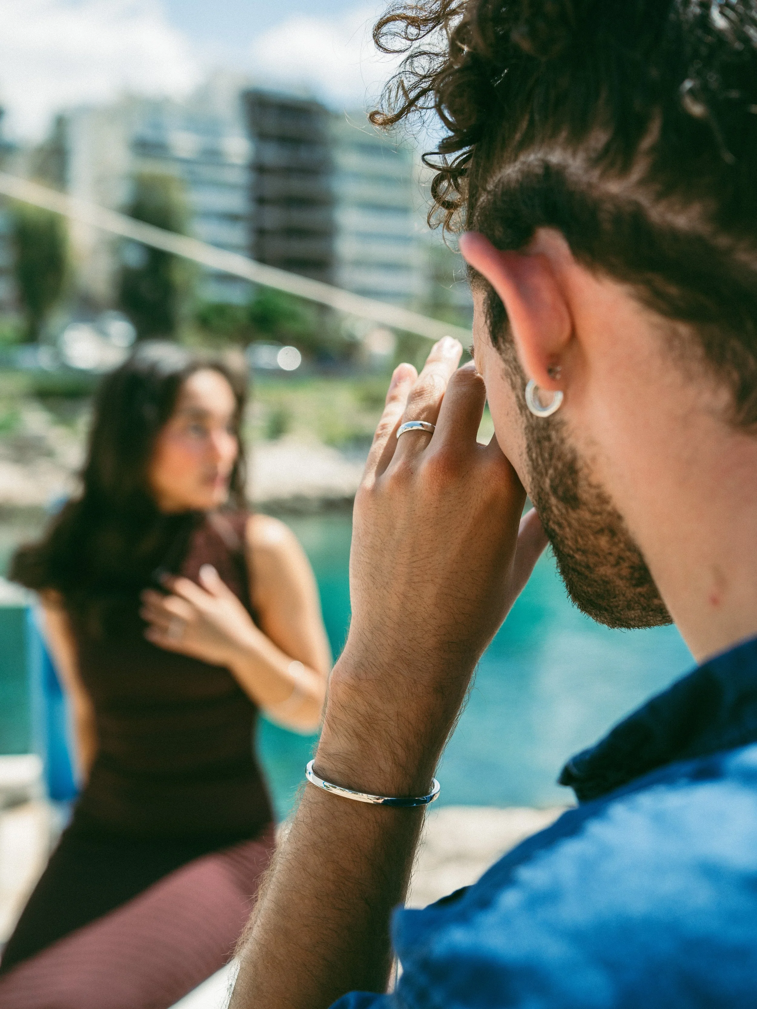 A man with curly hair and earrings is holding his hand over his face, looking at a woman in the background near water and buildings.