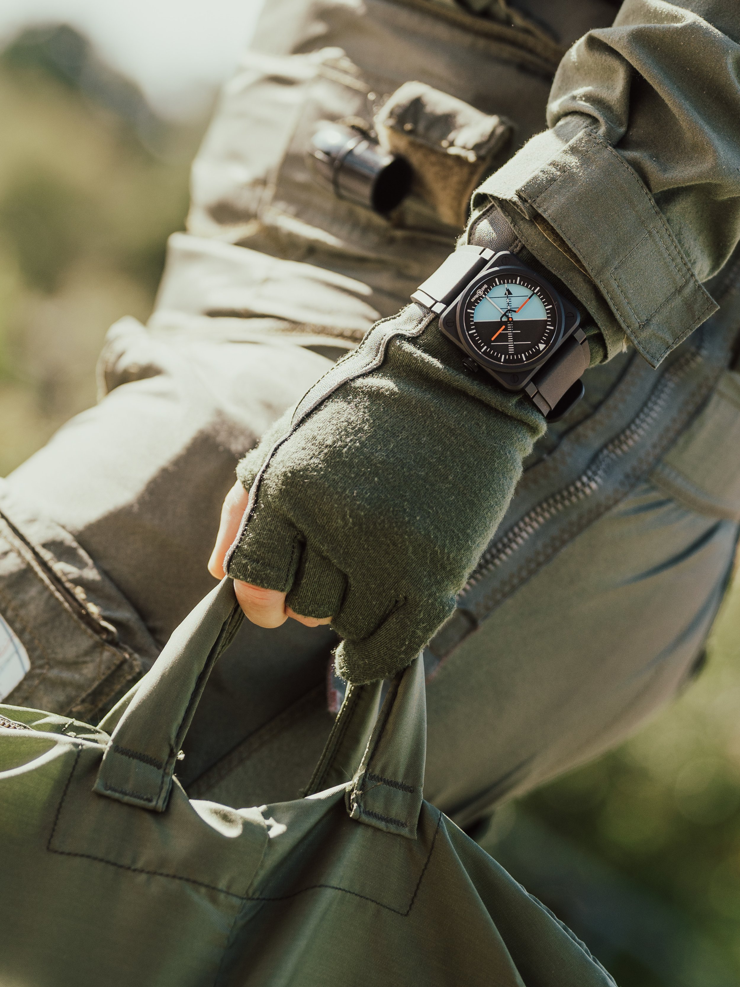 A person wearing tactical gear and a black smartwatch with a round face, holding a green bag with their gloved hand.