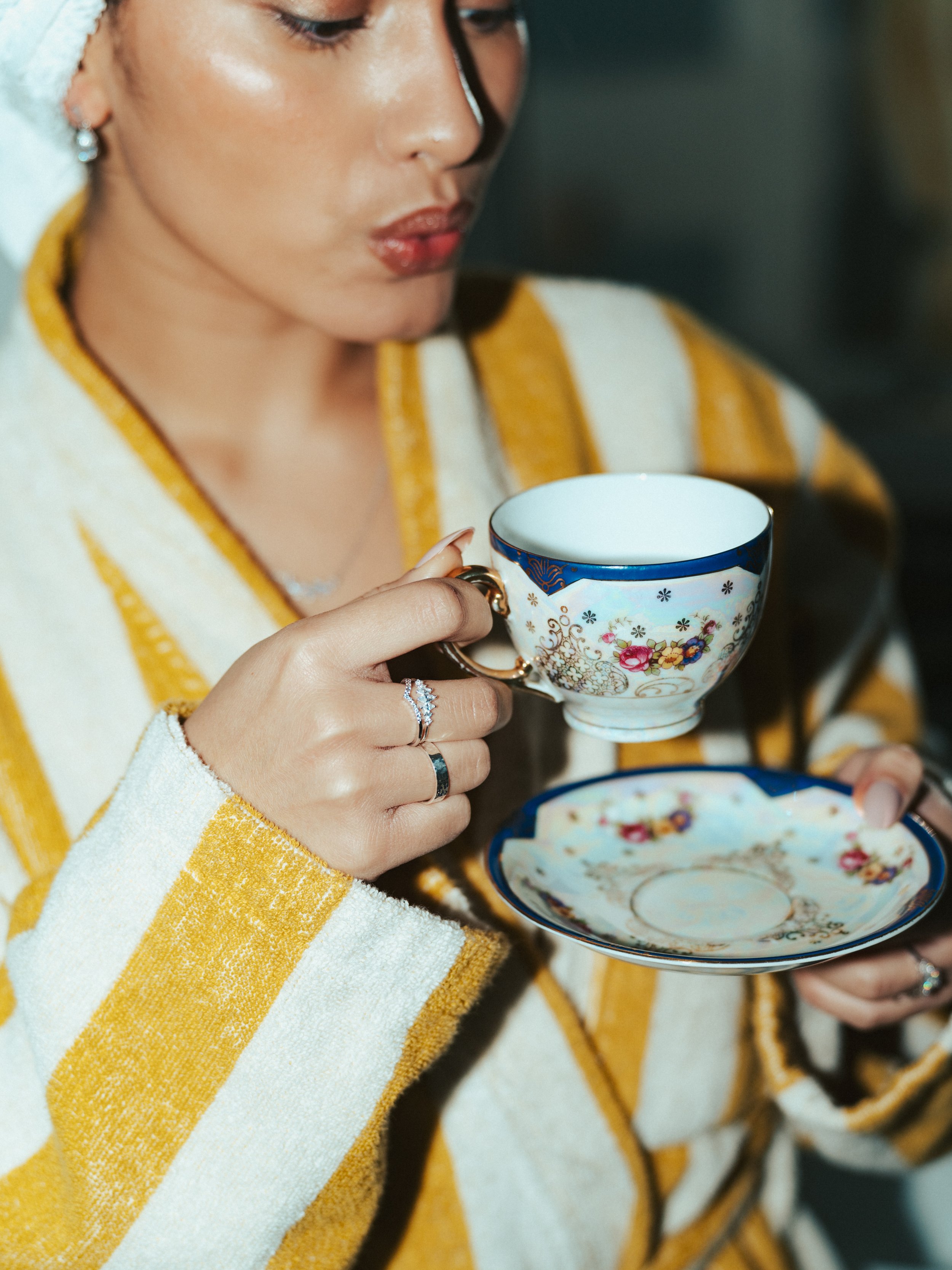 A woman with light skin is holding a floral-patterned teacup in one hand, with a matching saucer in her other hand. She is wearing a yellow and white striped robe and has silver jewelry, including rings and earrings.