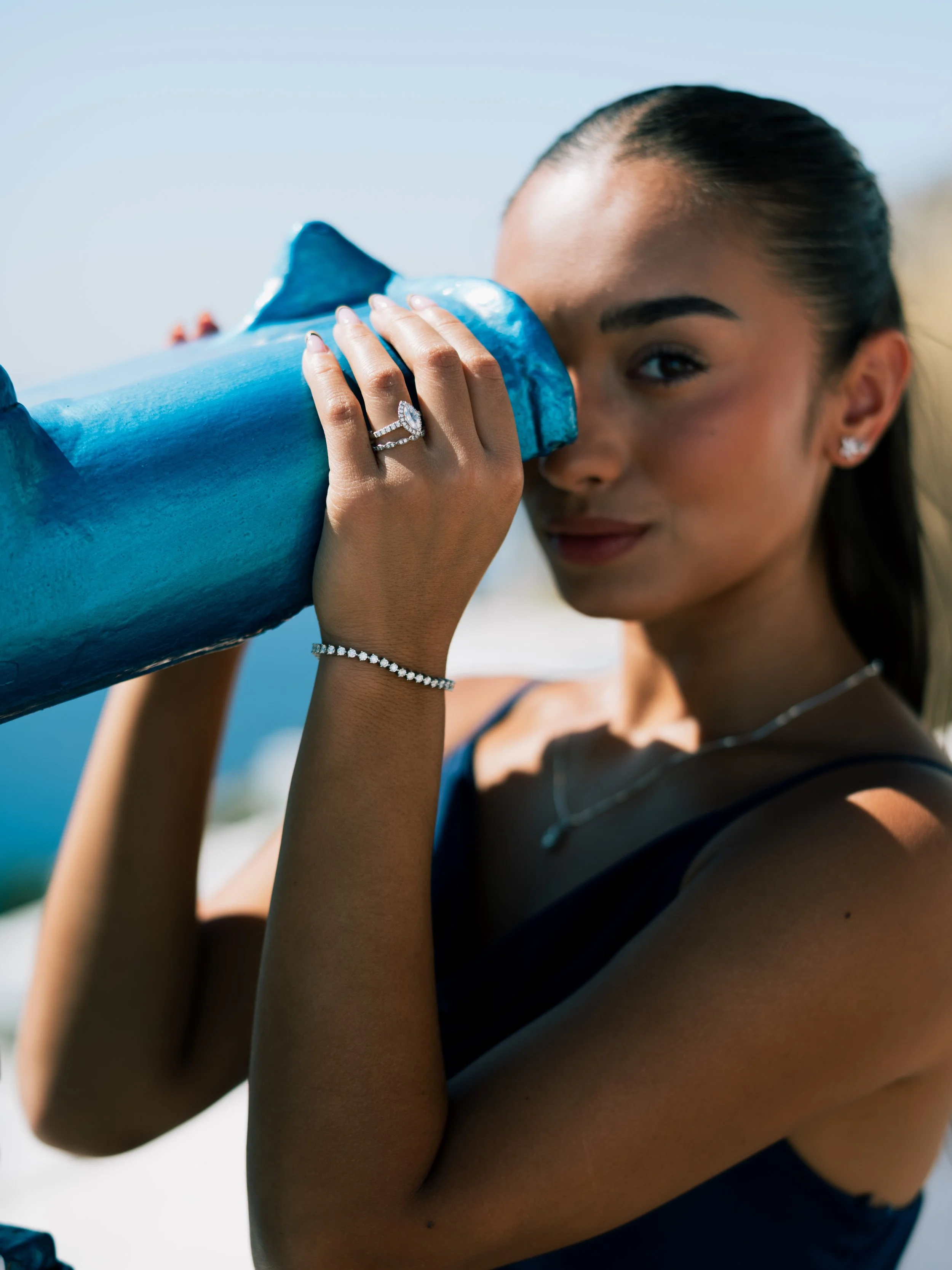 A woman with dark hair pulled back, wearing jewelry, looks through a blue telescope outdoors with a sky background.