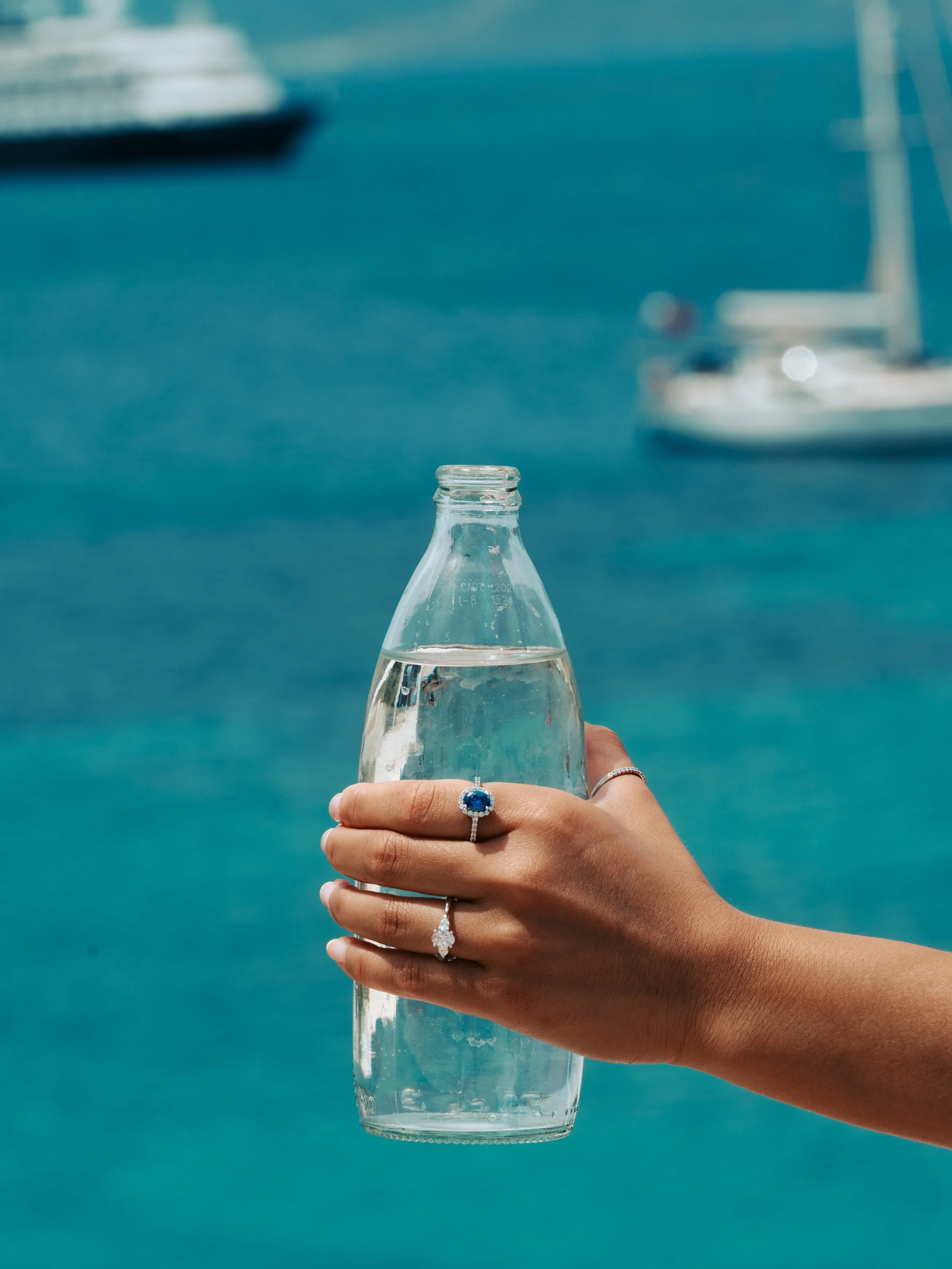 A hand with rings is holding a glass bottle of water against a background of blue water and boats.