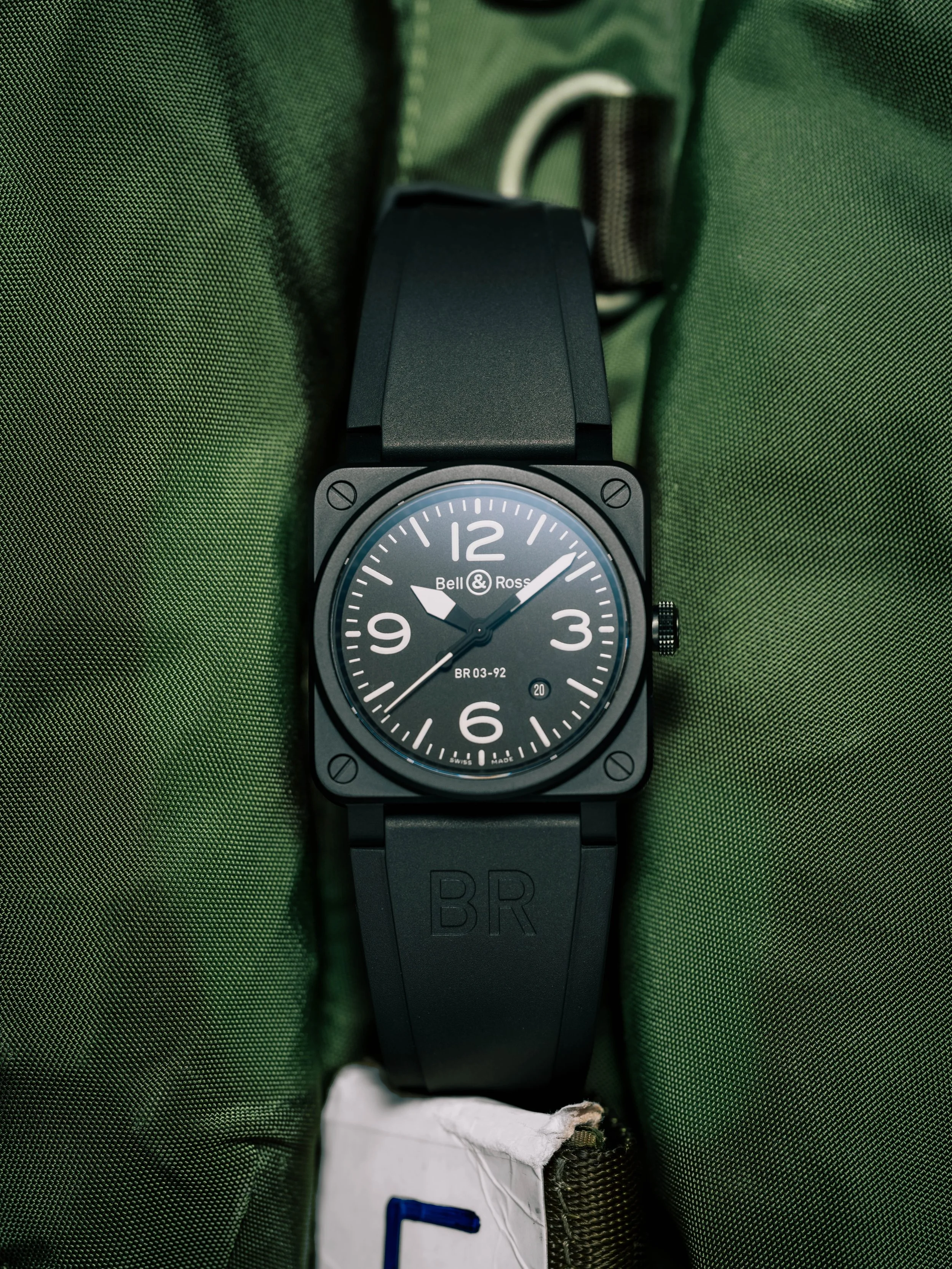 A black wristwatch with a square face and round dial showing the time, resting on a green fabric surface.
