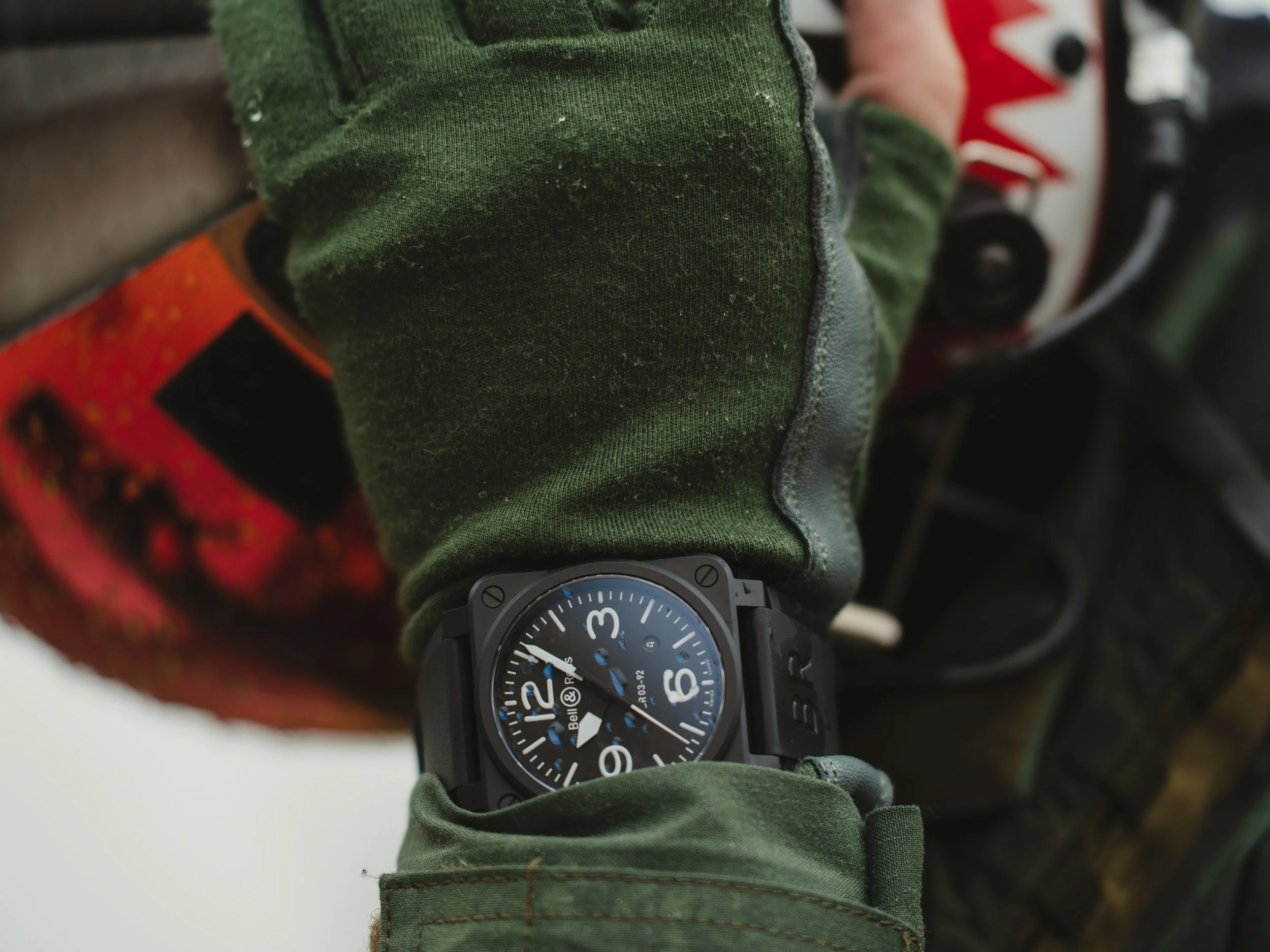 A person's wrist wearing a black Bell & Ross watch with a black dial and white numerals, over a green camouflage sleeve, with a motorcycle helmet and a part of a motorcycle in the background.