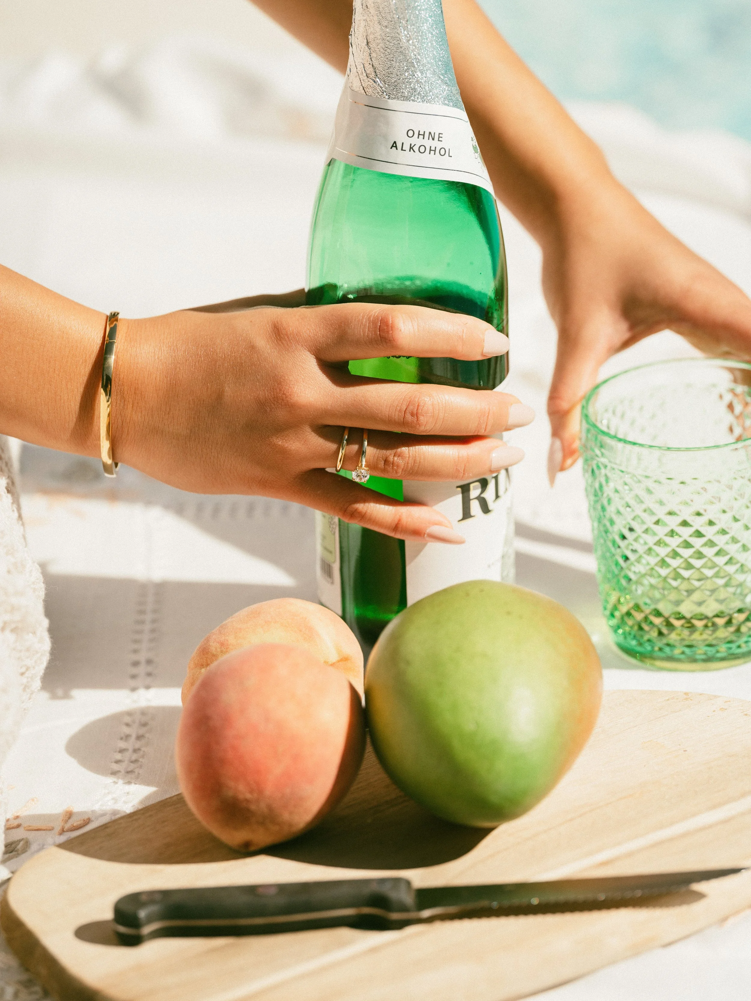 A person holding a green glass bottle in one hand and a textured green glass in the other, with peaches and an apple on a wooden cutting board, along with a black-handled knife, on a white tablecloth.