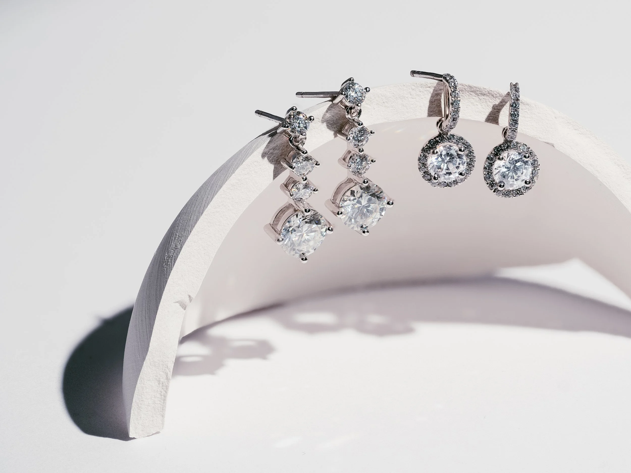 Set of three pairs of silver earrings with clear gemstones, displayed on a curved white surface against a white background.