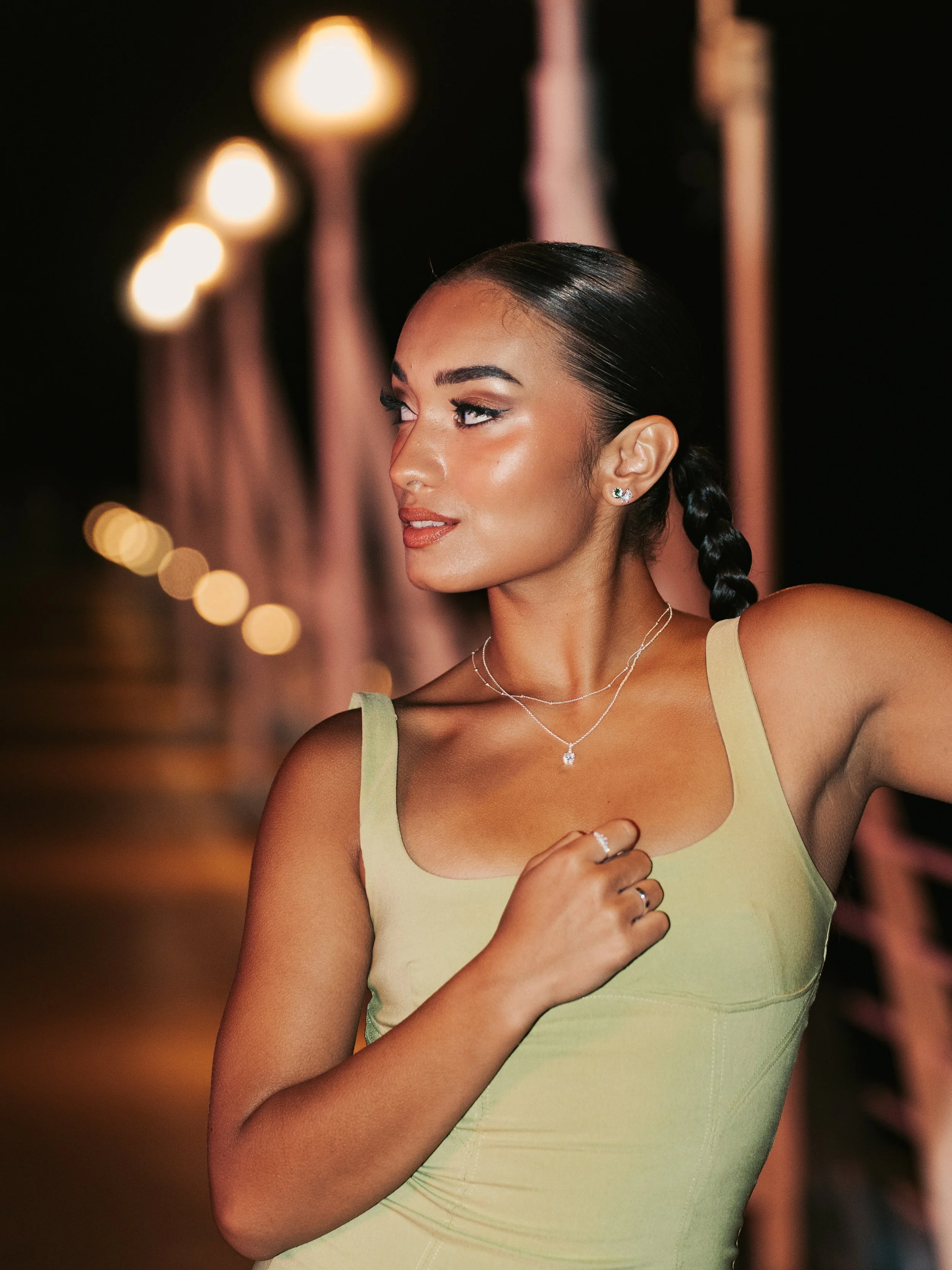 A woman at night with styled dark hair in a braid, wearing a light yellow sleeveless top and jewelry, standing near pole lights with blurred background.