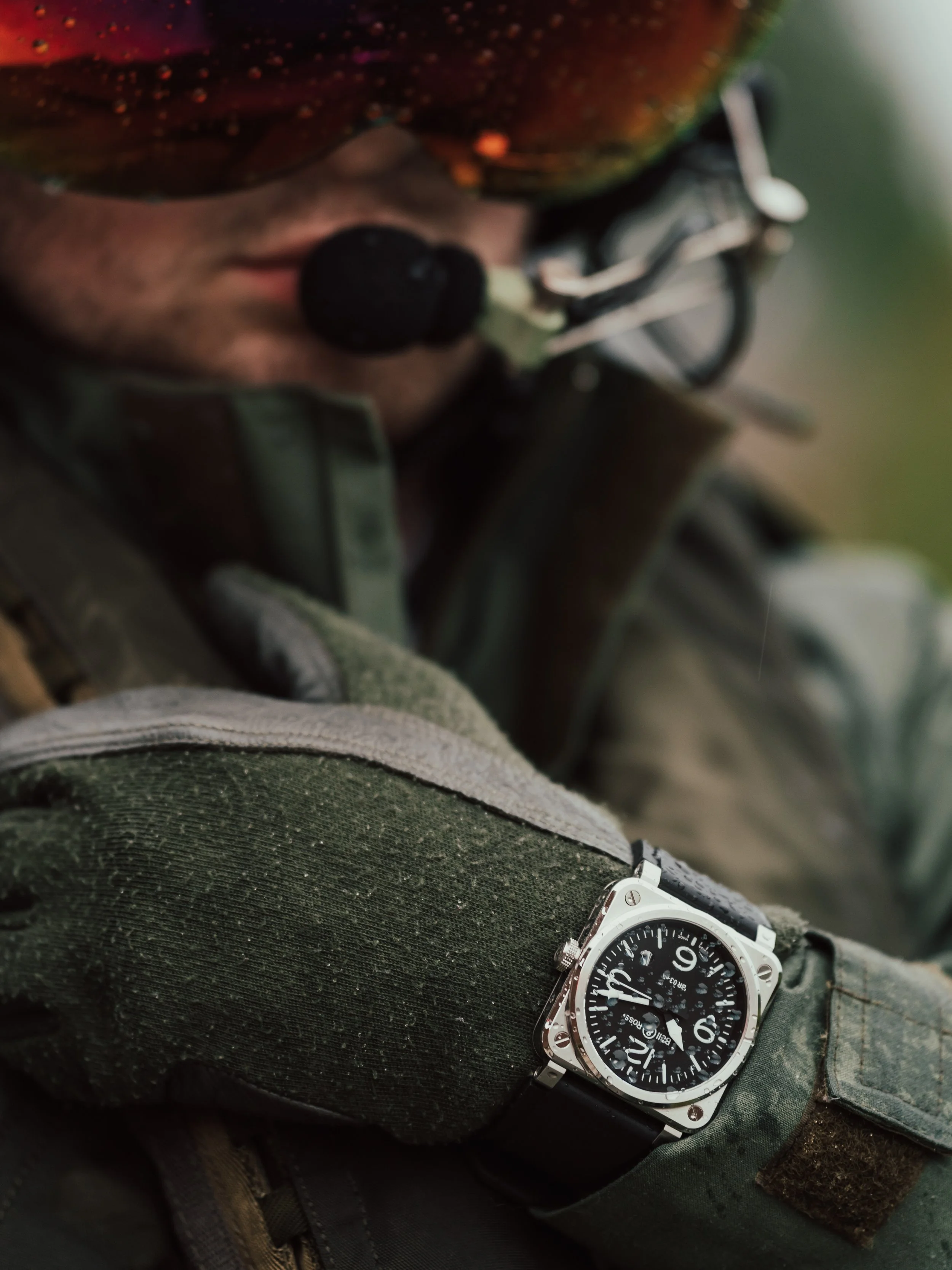 Close-up of a soldier's wrist wearing a large, black dial Bell & Ross square watch with a black strap, with a soldier in the background wearing a camouflage helmet and uniform, focusing on a radio microphone near his mouth.