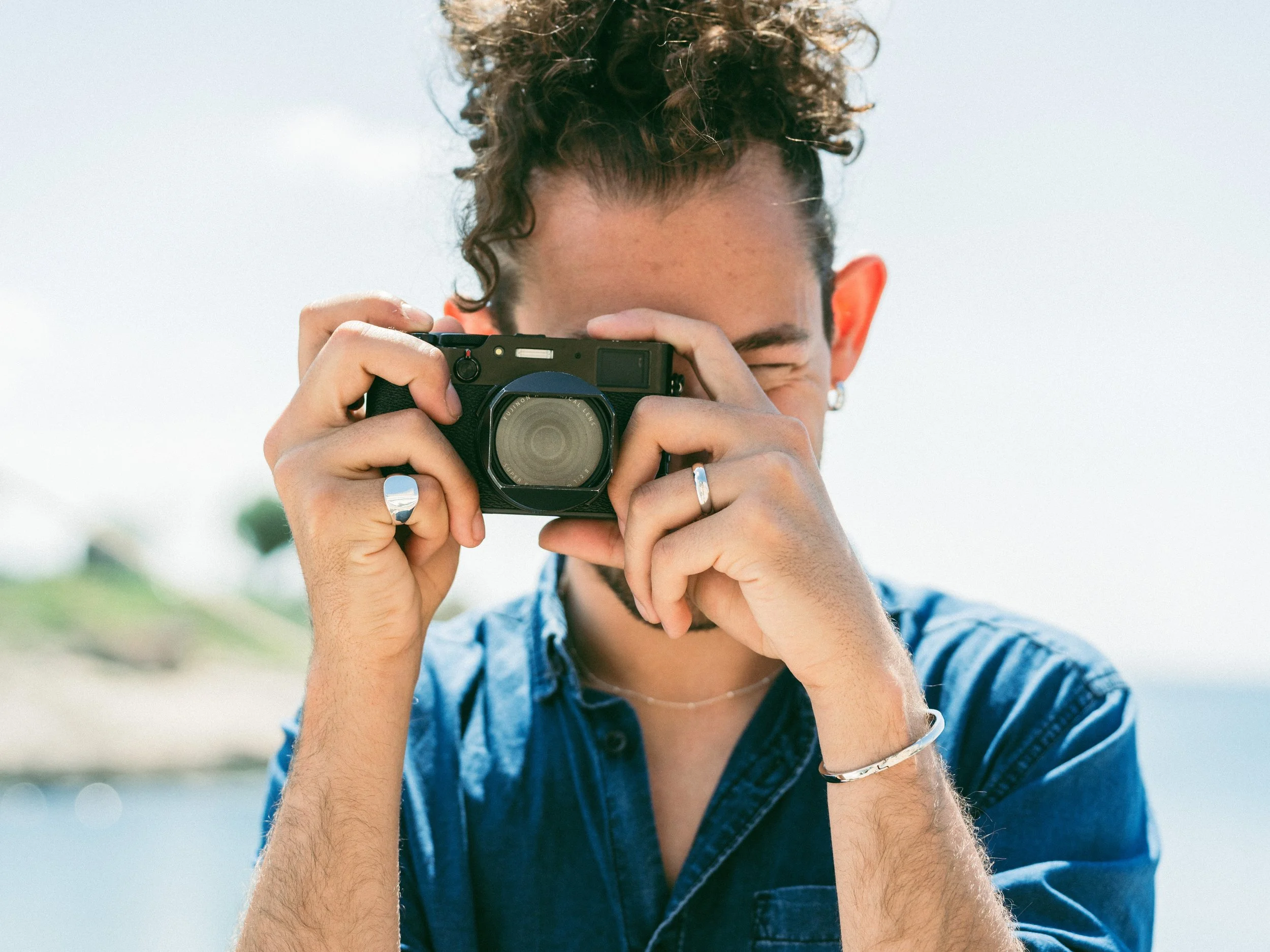 Person with curly hair taking a photo with a camera outdoors on a bright day.