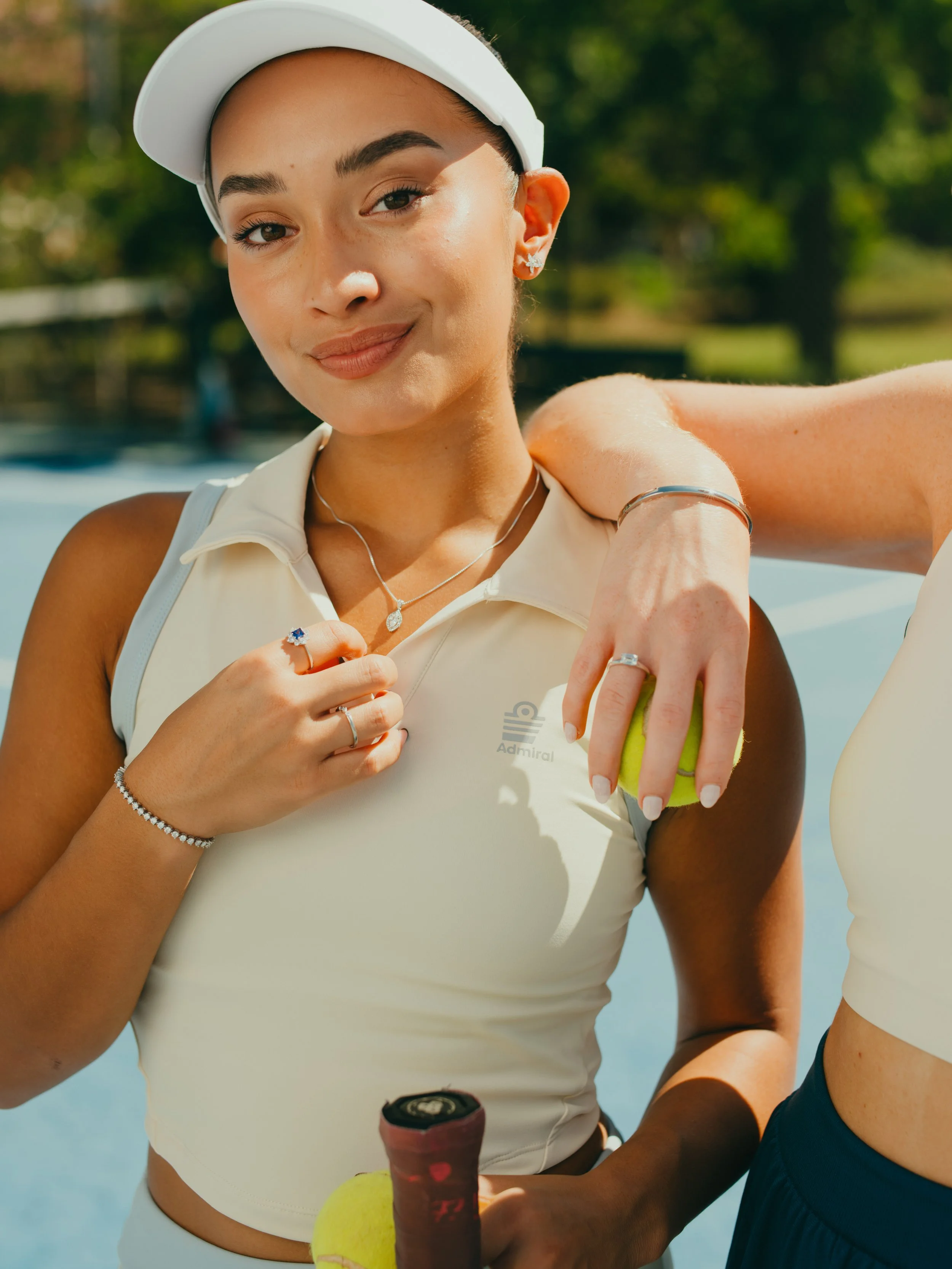 Young woman in athletic wear with jewelry, smiling outdoors near water, holding a tennis ball and a tennis racket.