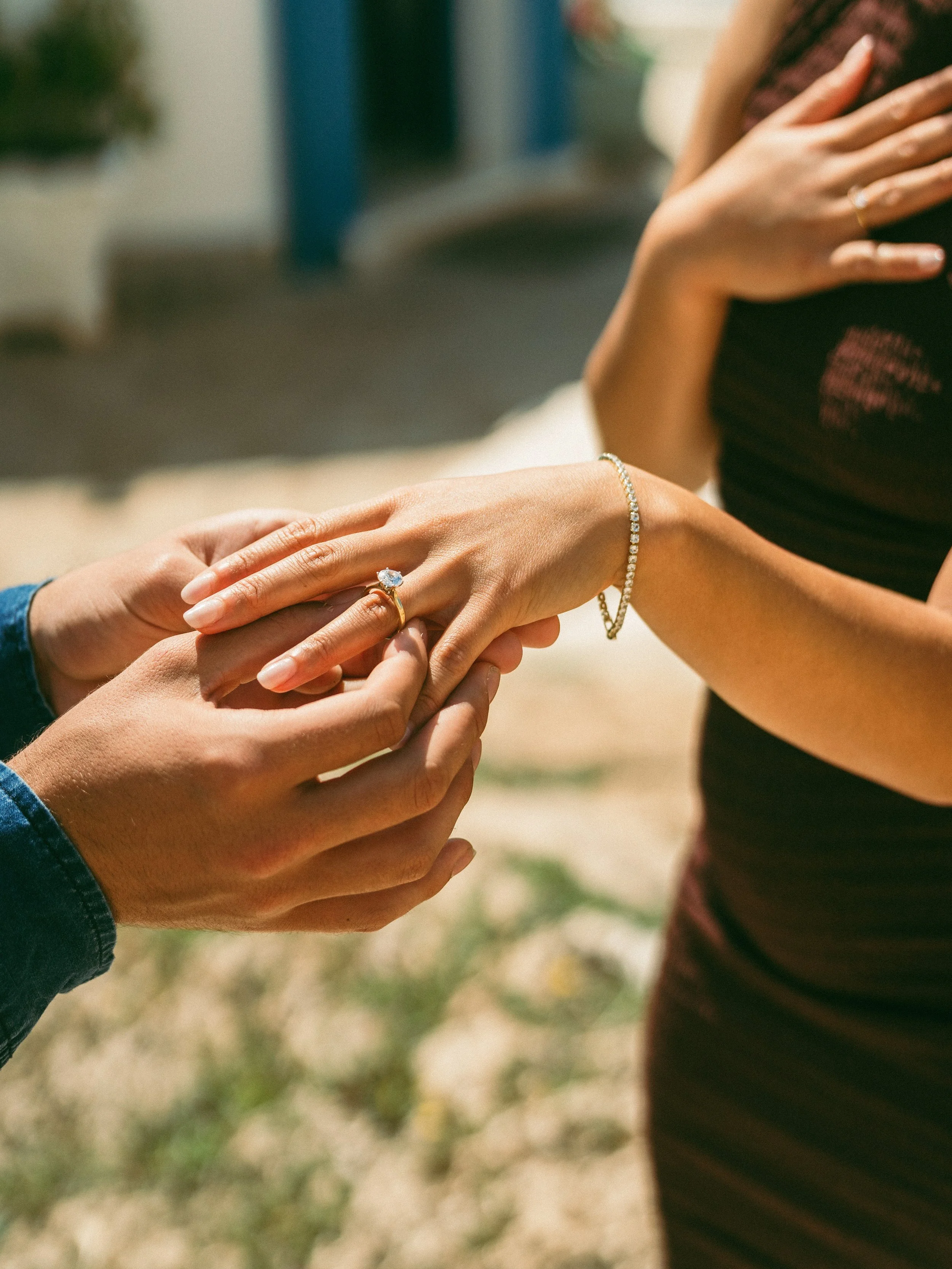 Person is getting engaged outdoors, with the person on the right receiving the ring and the person on the left placing it on her finger.