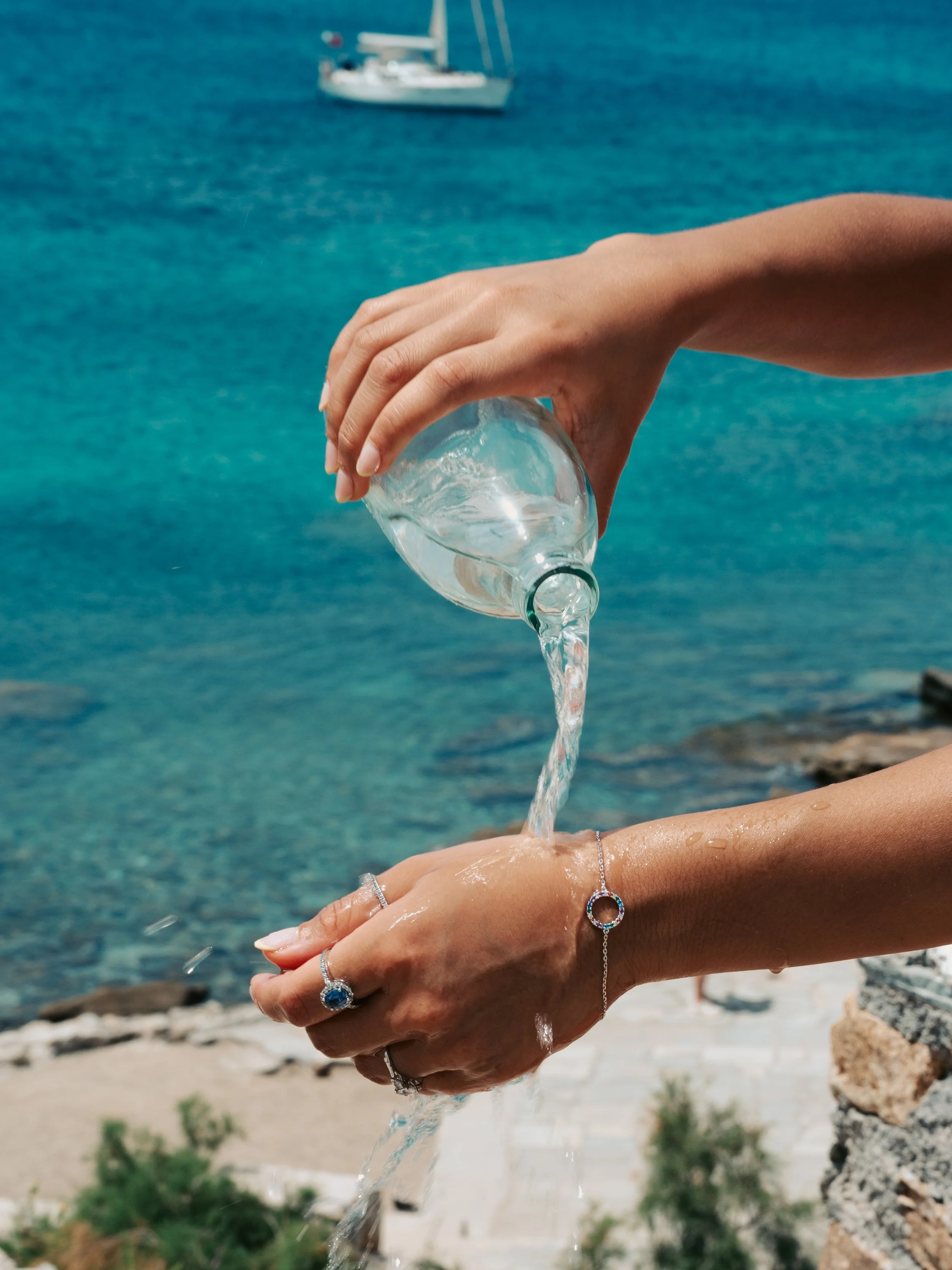 Hands holding a glass bottle pouring water over wrist adorned with jewelry, set against a background of the ocean with a boat in the distance.