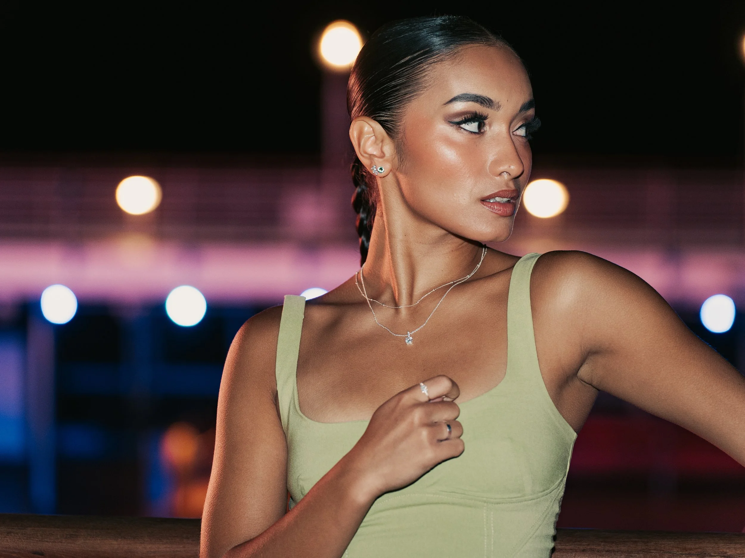 A woman with dark hair styled in a braid, wearing a green sleeveless top, jewelry, and makeup, looking to the side with a nighttime city background of blurred lights.