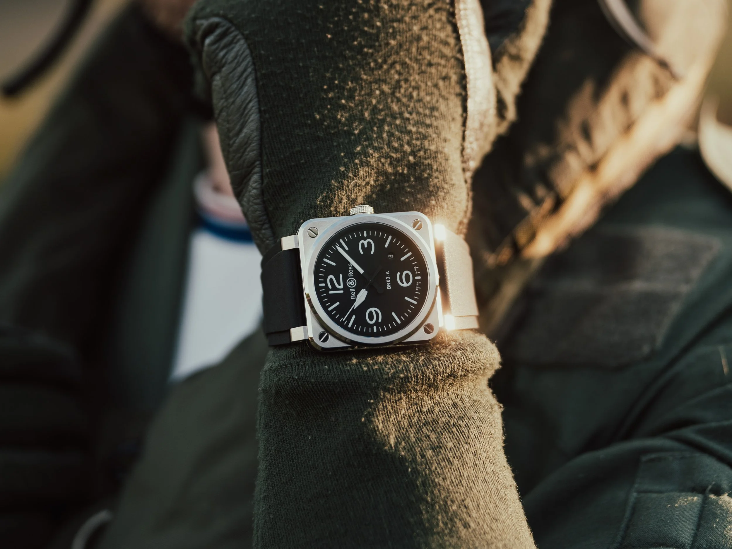 Close-up of a wrist wearing a black and silver watch with a black dial, showing the time, on a person's arm covered with a dark glove, outdoor setting with sunlight.