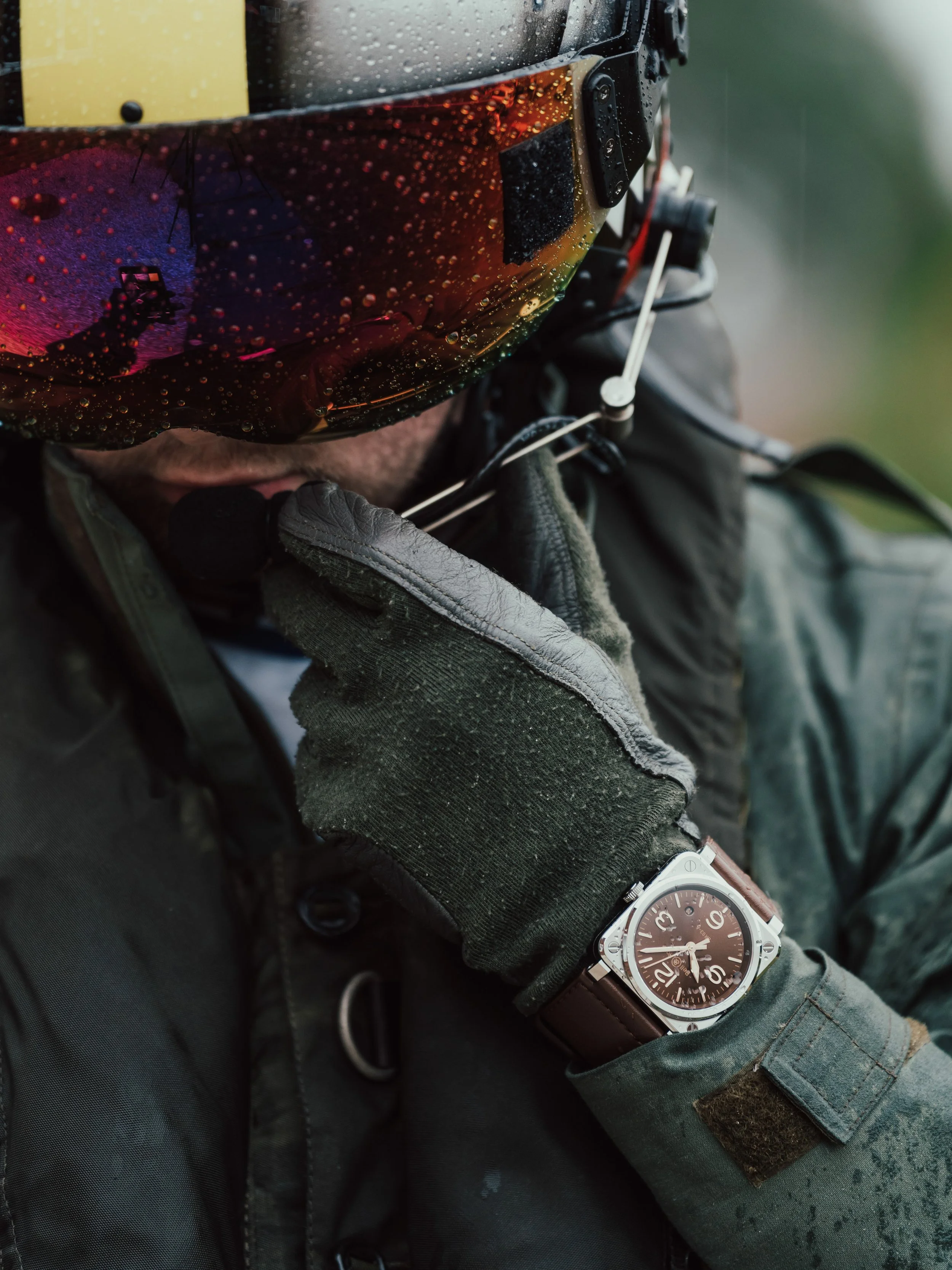 Close-up of a person wearing a black helmet with water droplets, green gloves, and a military-style jacket, adjusting their gear, with a watch visible on their wrist.