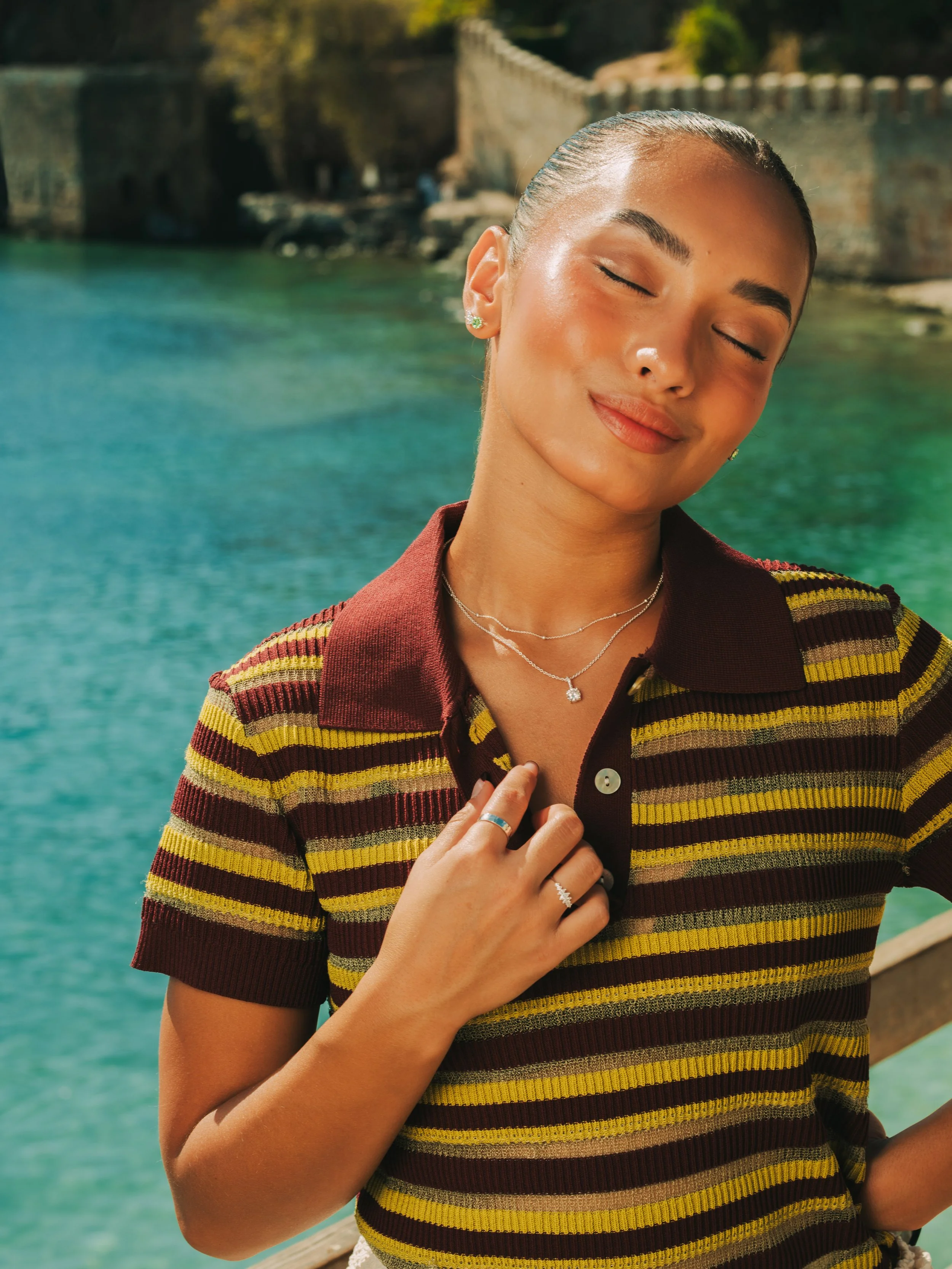 Woman with her eyes closed and a gentle smile, wearing a striped polo shirt and jewelry, standing near a river with a bridge in the background.