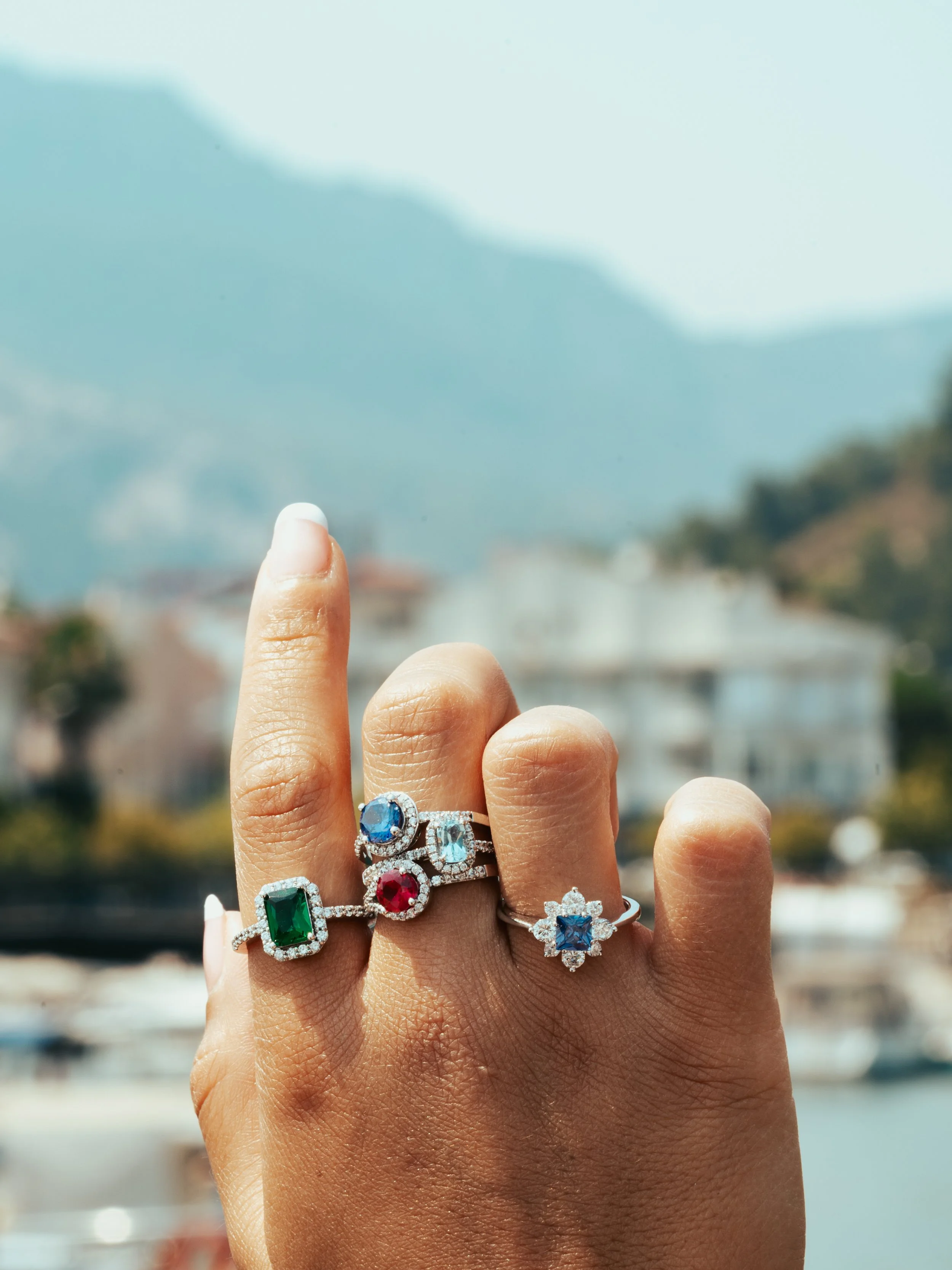 A person's hand showcasing multiple rings with colorful gemstones, set against a blurred outdoor background with mountains and buildings.