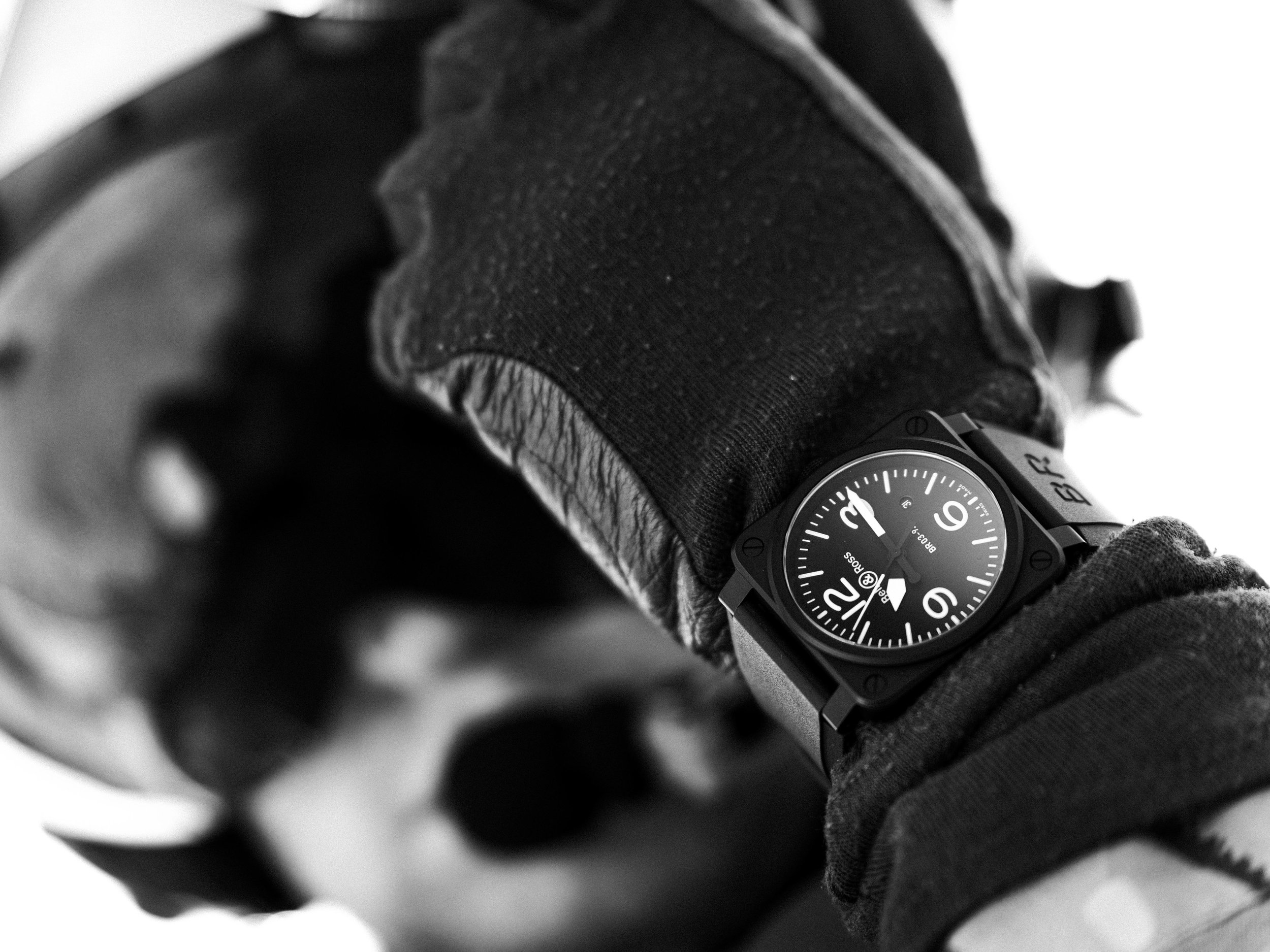 A close-up black-and-white image of a person's wrist wearing a black watch with a white dial, worn over a sleeve of a black glove.