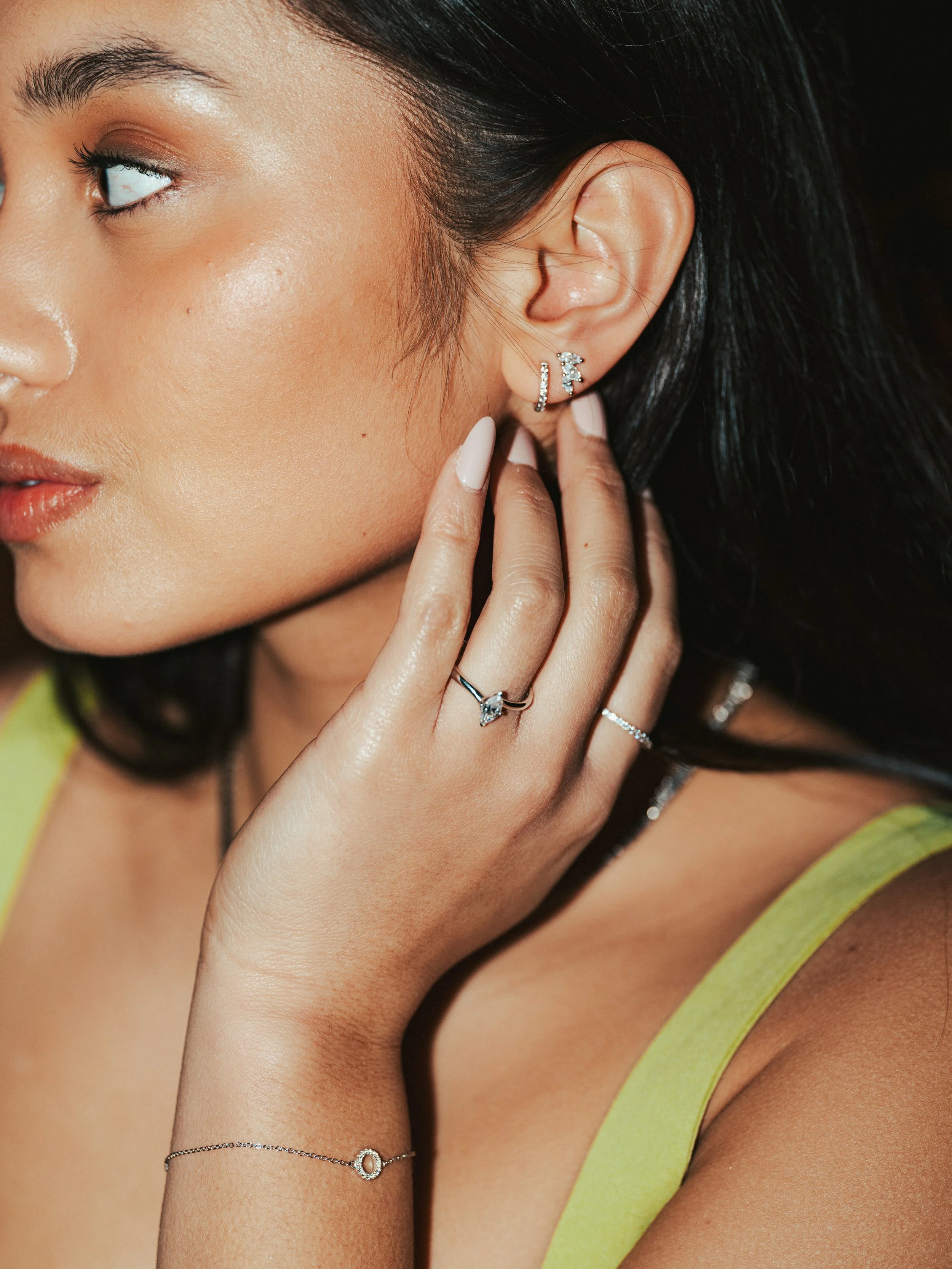 Close-up of a woman's face and hand showing jewelry, including earrings, rings, and a bracelet, with a yellow top.