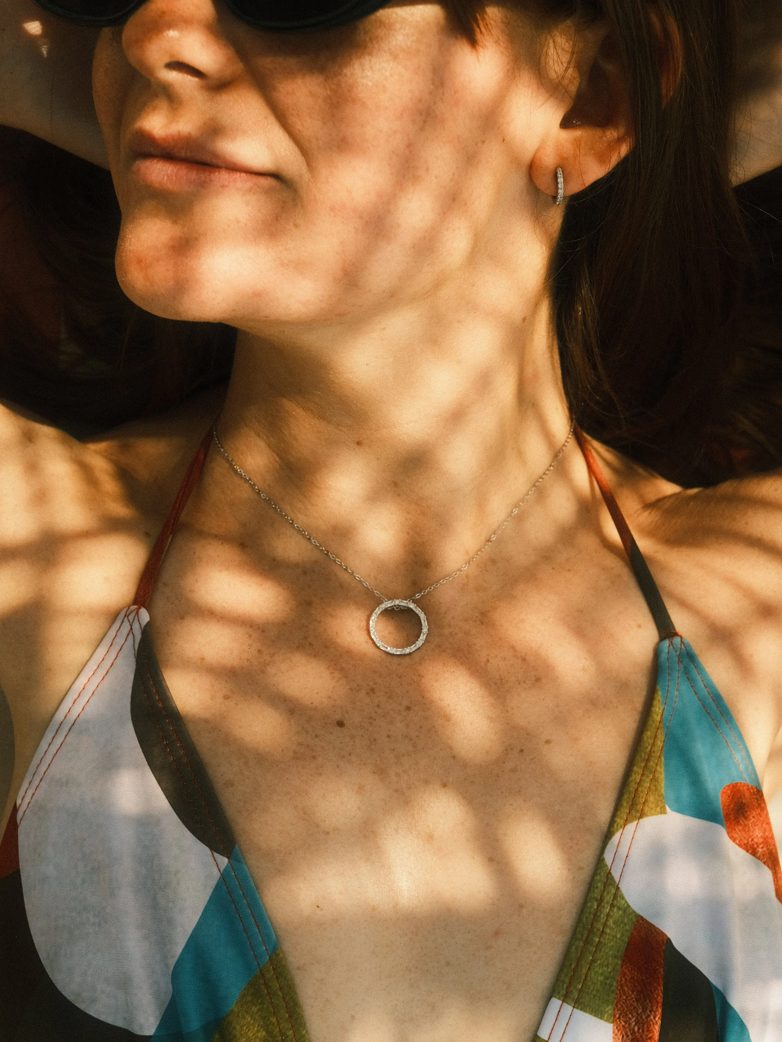 Close-up of a woman lying down, wearing sunglasses, a colorful patterned top, and jewelry including a circular pendant necklace and a hoop earring, with shadows of leaves on her skin.