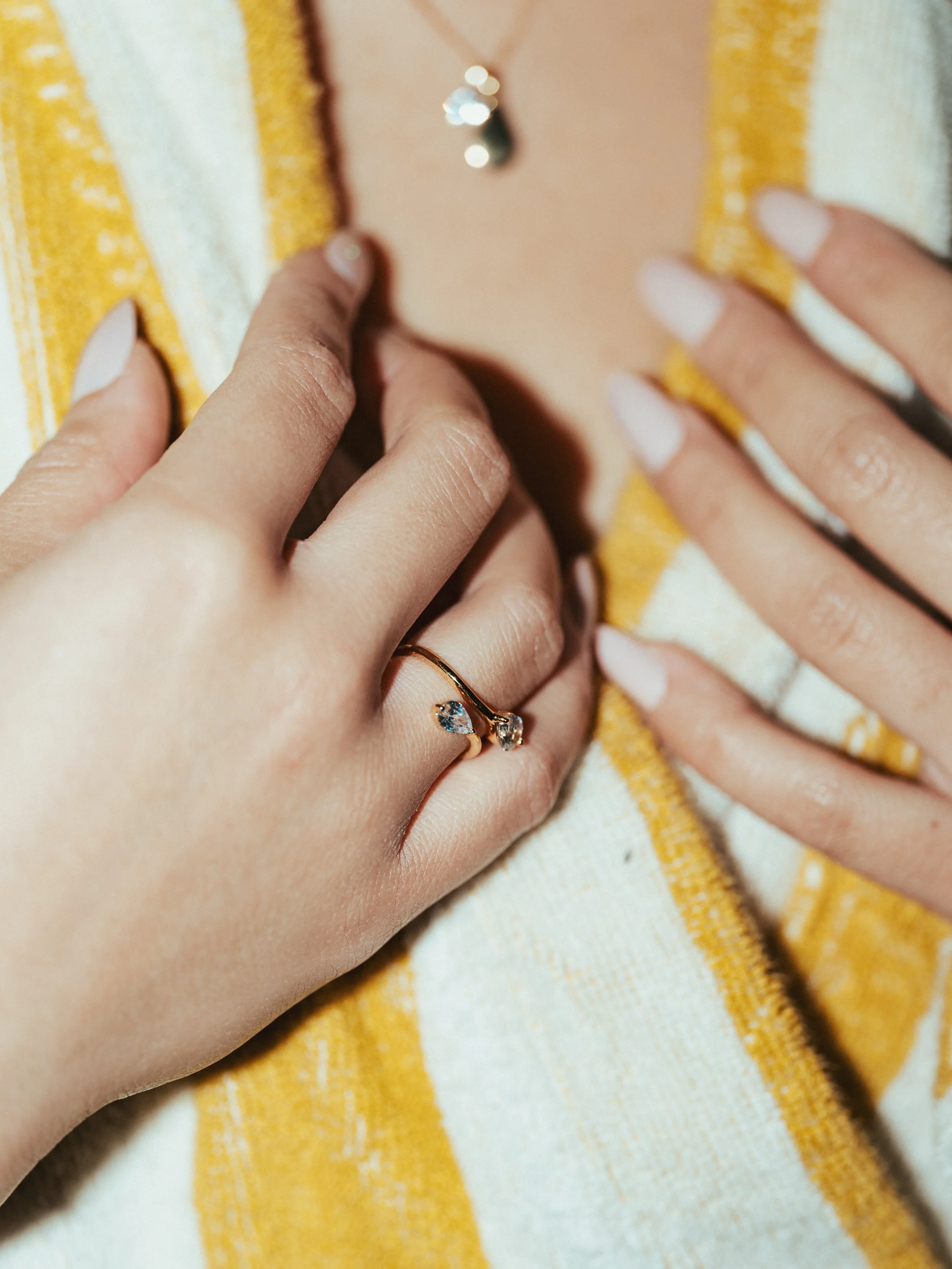 Close-up of a woman's hand wearing a gold ring with a small diamond, touching her necklace, with a partial view of her chest and fingers with French manicure, against a yellow and white striped fabric background.