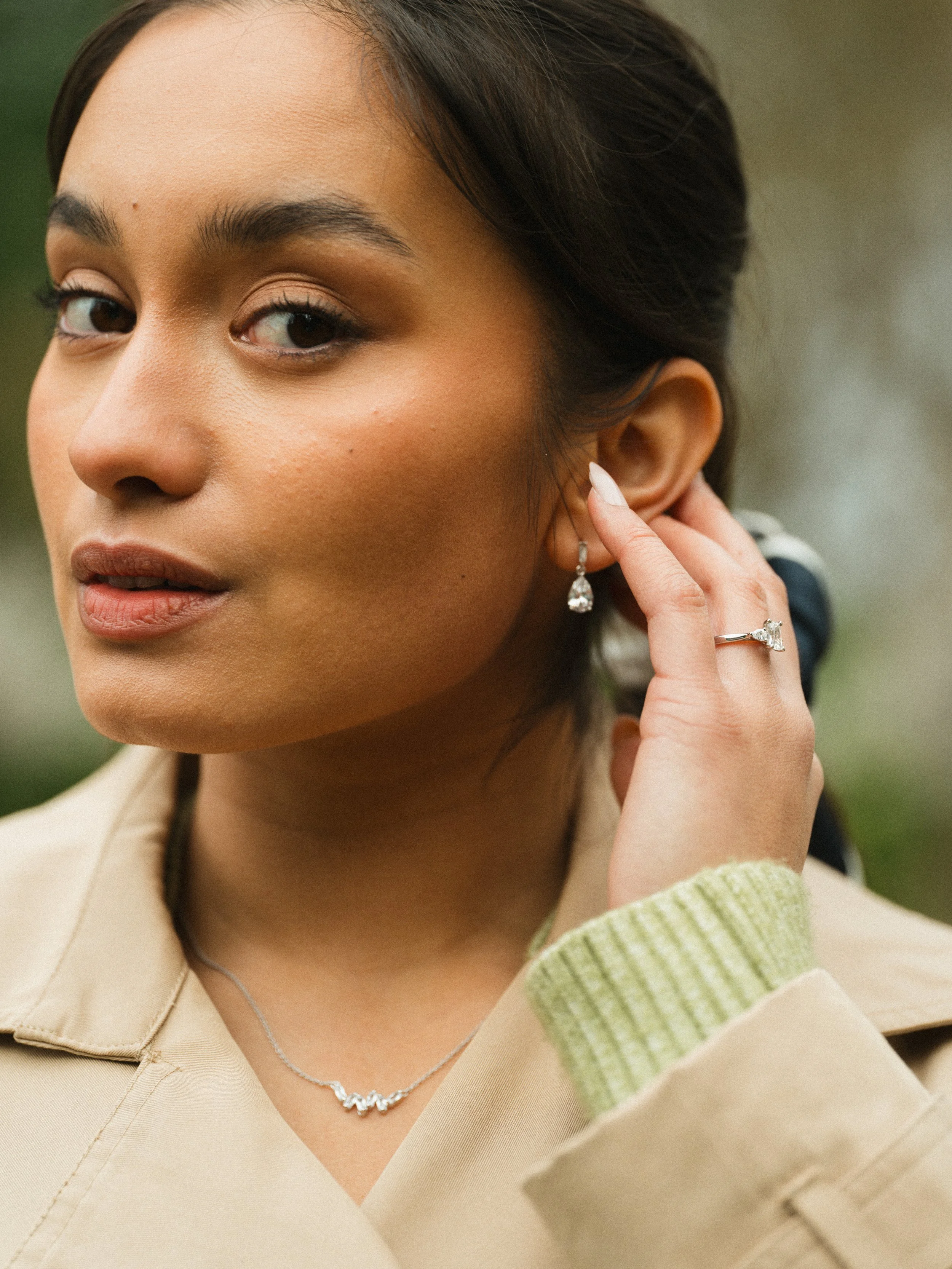 A woman with dark hair styled back, wearing earrings, a necklace, a ring, and a beige coat with a light green ribbed cuff, adjusting her earring. The background is blurred greenery.