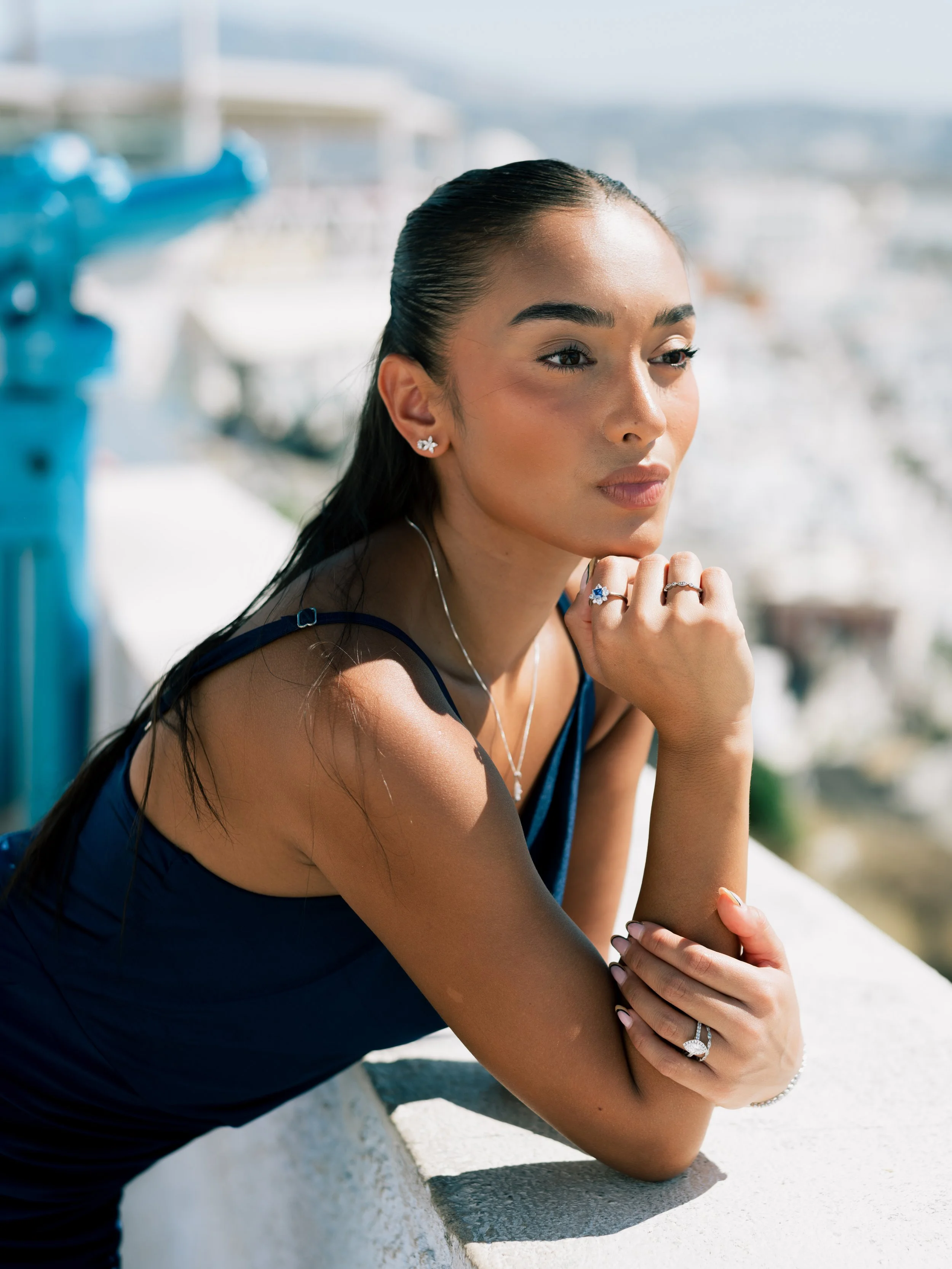 A woman with dark hair tied back, wearing jewelry including earrings and rings, leans on a white ledge outdoors with boats and water in the background.