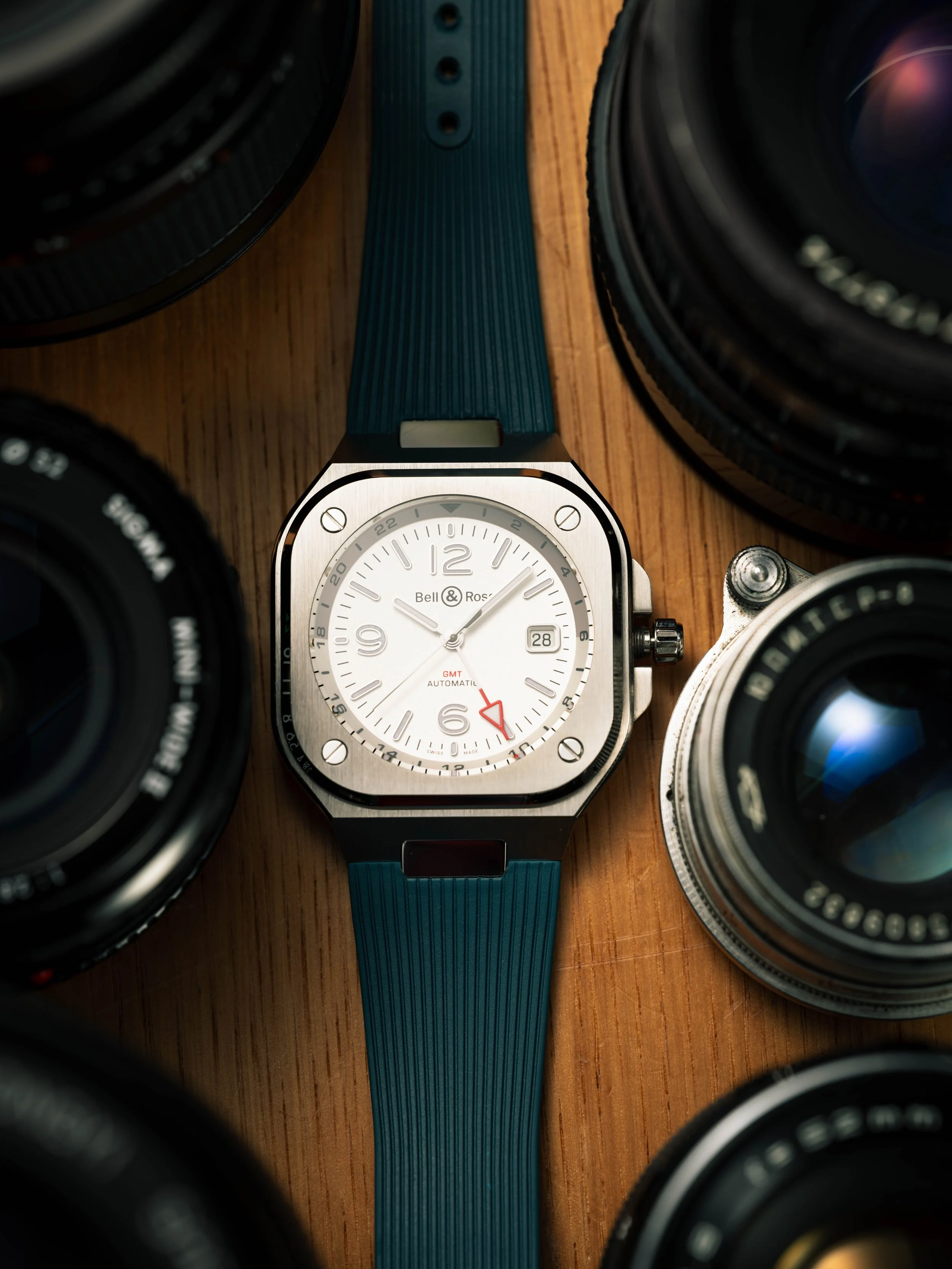 A luxury watch with a white face, black and silver casing, and a blue rubber strap, surrounded by several camera lenses on a wooden surface.
