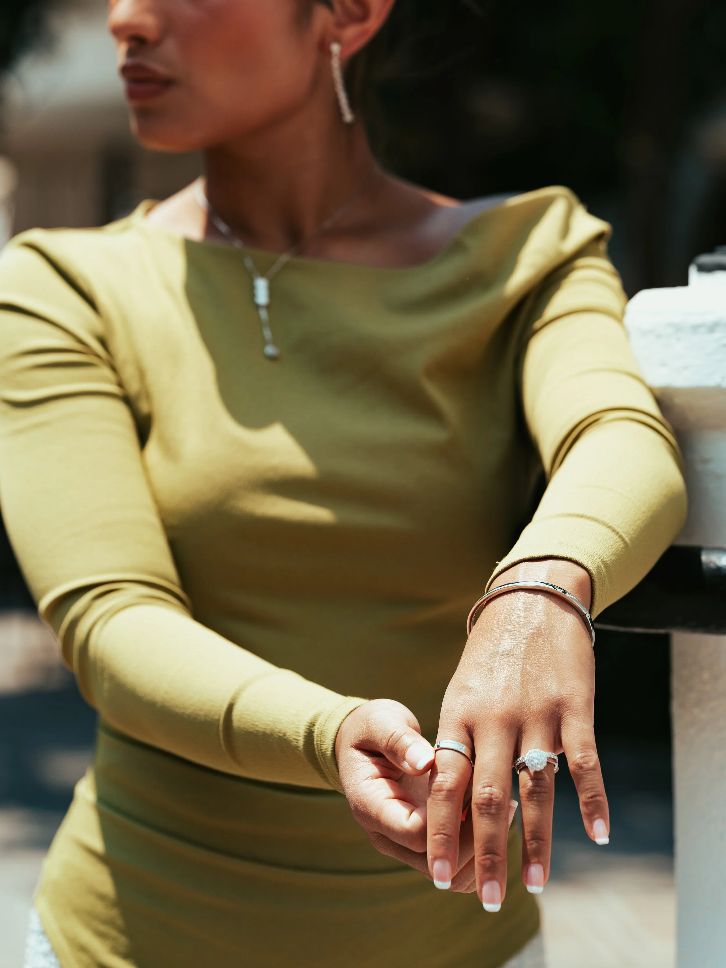 Close-up of a woman wearing a yellow long-sleeve top, showing her hand with diamond rings and bracelets, and jewelry including earrings, a necklace, and a bracelet.