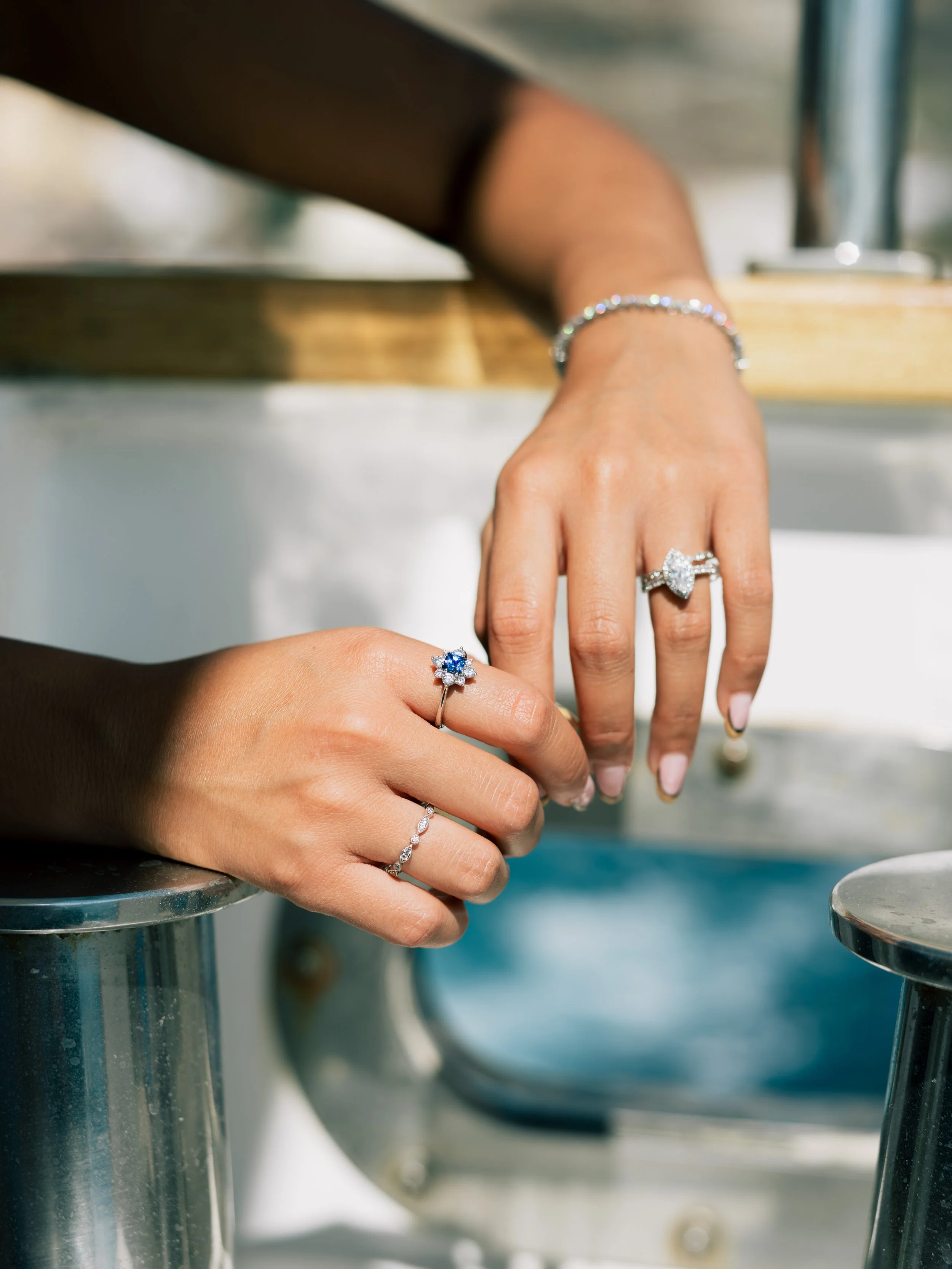 Two women showcase their hands with sparkling rings, one with a blue gemstone and the other with a large clear stone, and jewelry including a bracelet and a delicate diamond ring, sitting near a yacht or boat.