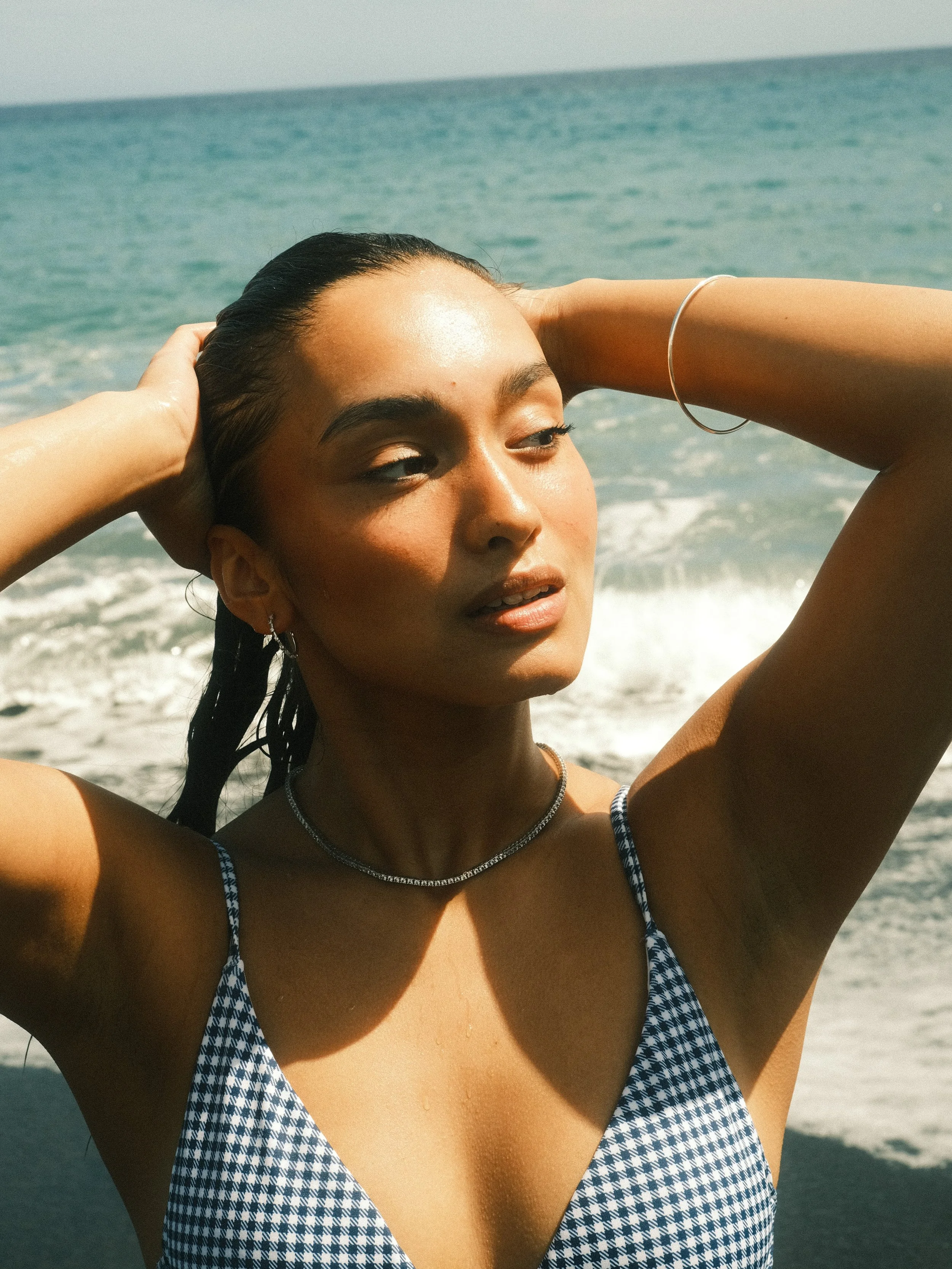 A woman with wet hair standing on the beach with her hands behind her head, overlooking the ocean, wearing a blue and white patterned swimsuit.