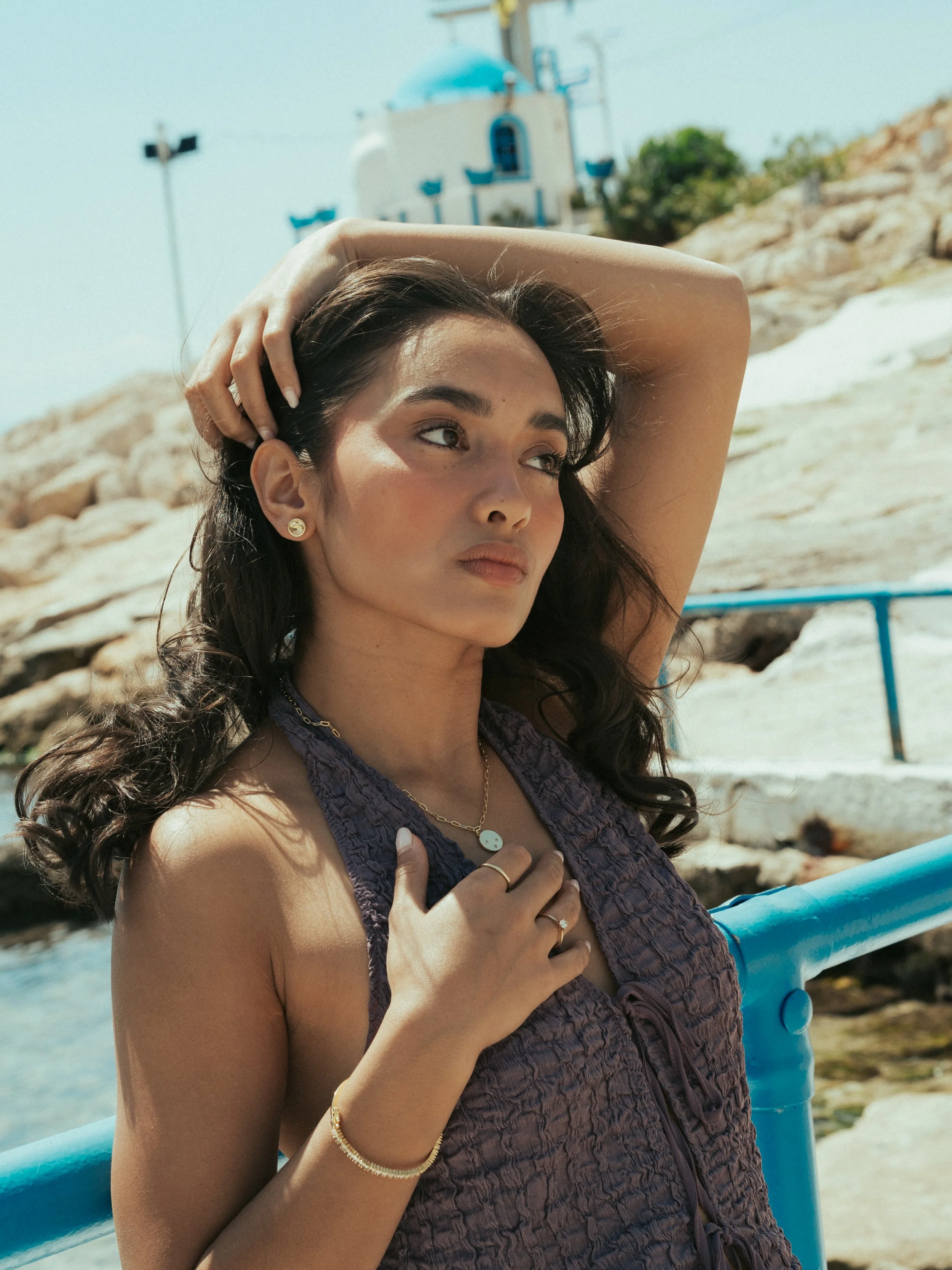 Young woman with dark wavy hair and jewelry posing outdoors near a body of water, with white buildings and rocks in the background.