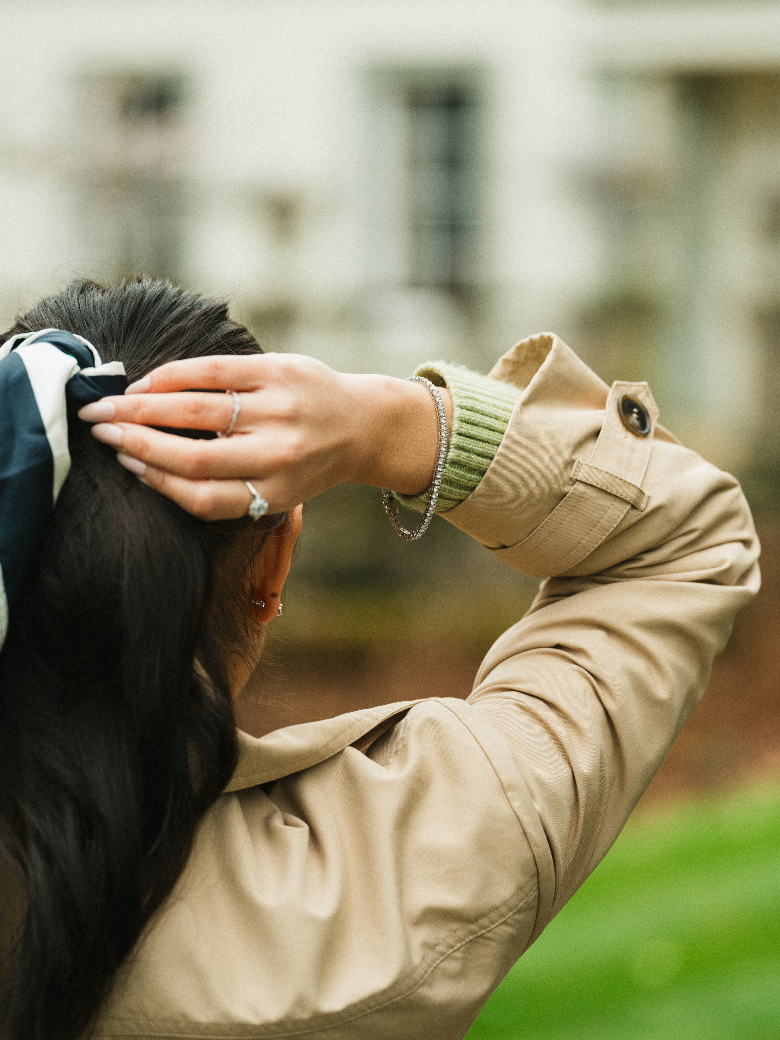 Person with long dark hair, wearing a beige trench coat and a green sweater, holding their head with both hands, standing outdoors with blurred buildings in the background.