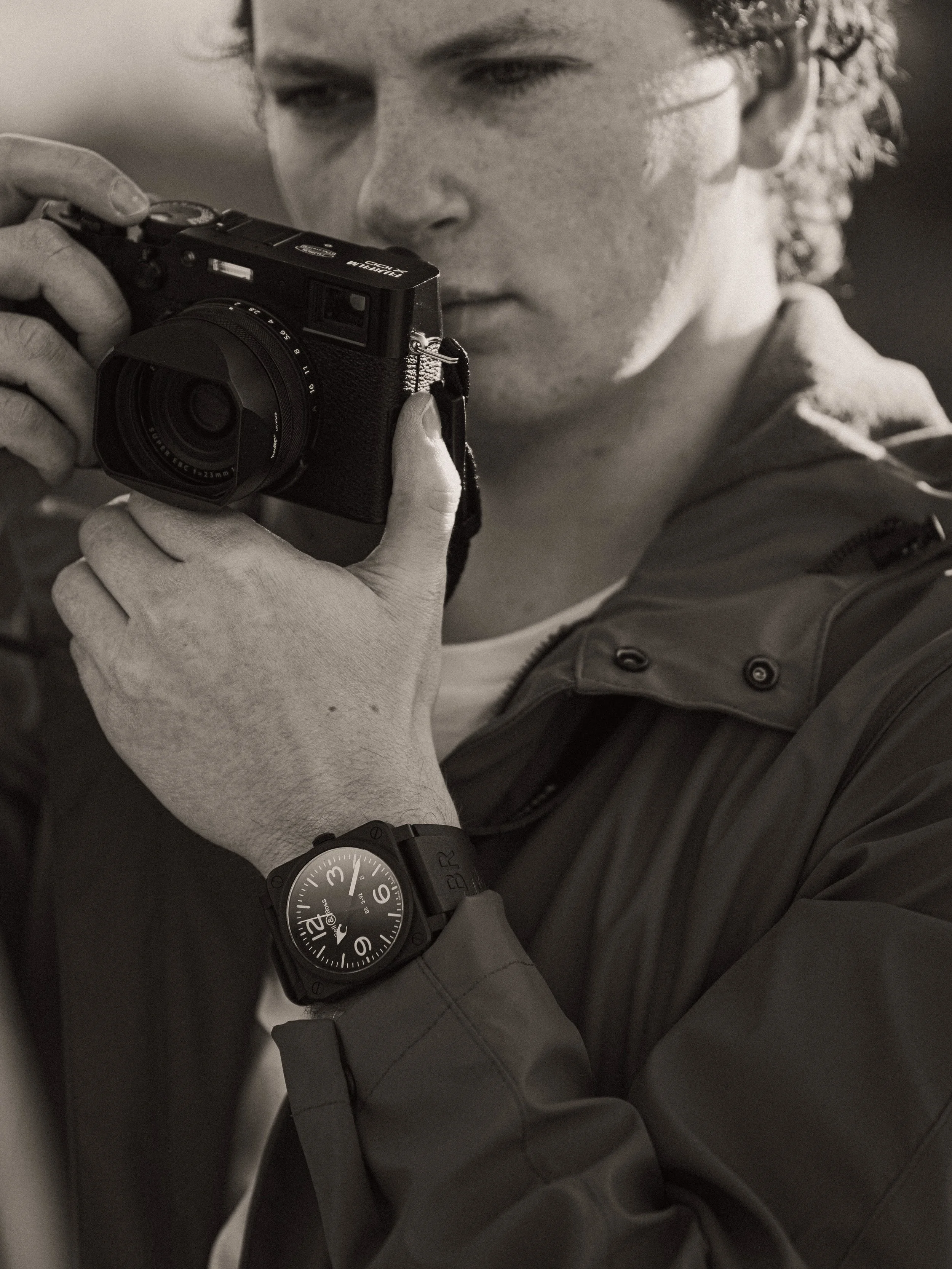 A person with curly hair, wearing a watch, looking through the viewfinder of a camera, dressed in a jacket and a t-shirt.