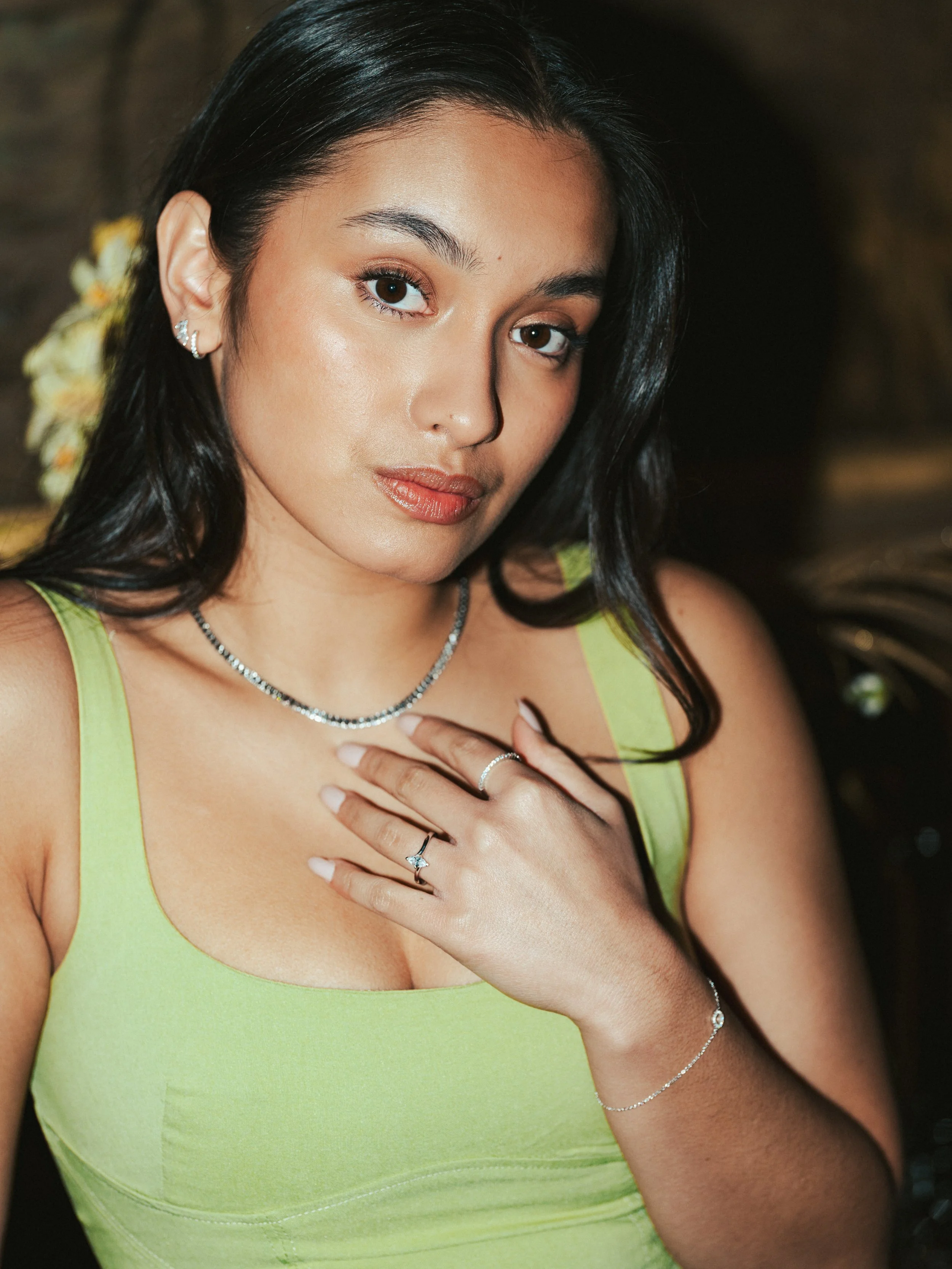 A young woman with long black hair and brown eyes wearing a lime green sleeveless top, jewelry including rings, earrings, a necklace, and a bracelet, touching her chest with her left hand. She is in an indoor setting with a blurred background.